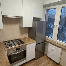 Warmińskie Domy PATRYK GICZEWSKI - Jasna kuchnia z drewnianą podłogą i białymi szafkami. Widoczna kuchenka, piekarnik i lodówka. Okno z widokiem na drzewa. Minimalistyczny design.