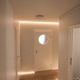 Warmińskie Domy PATRYK GICZEWSKI - Biały korytarz z drewnianą podłogą i oświetleniem LED przy suficie i podłodze. Na wprost drzwi z okrągłym oknem. Po bokach drzwi do innych pomieszczeń.