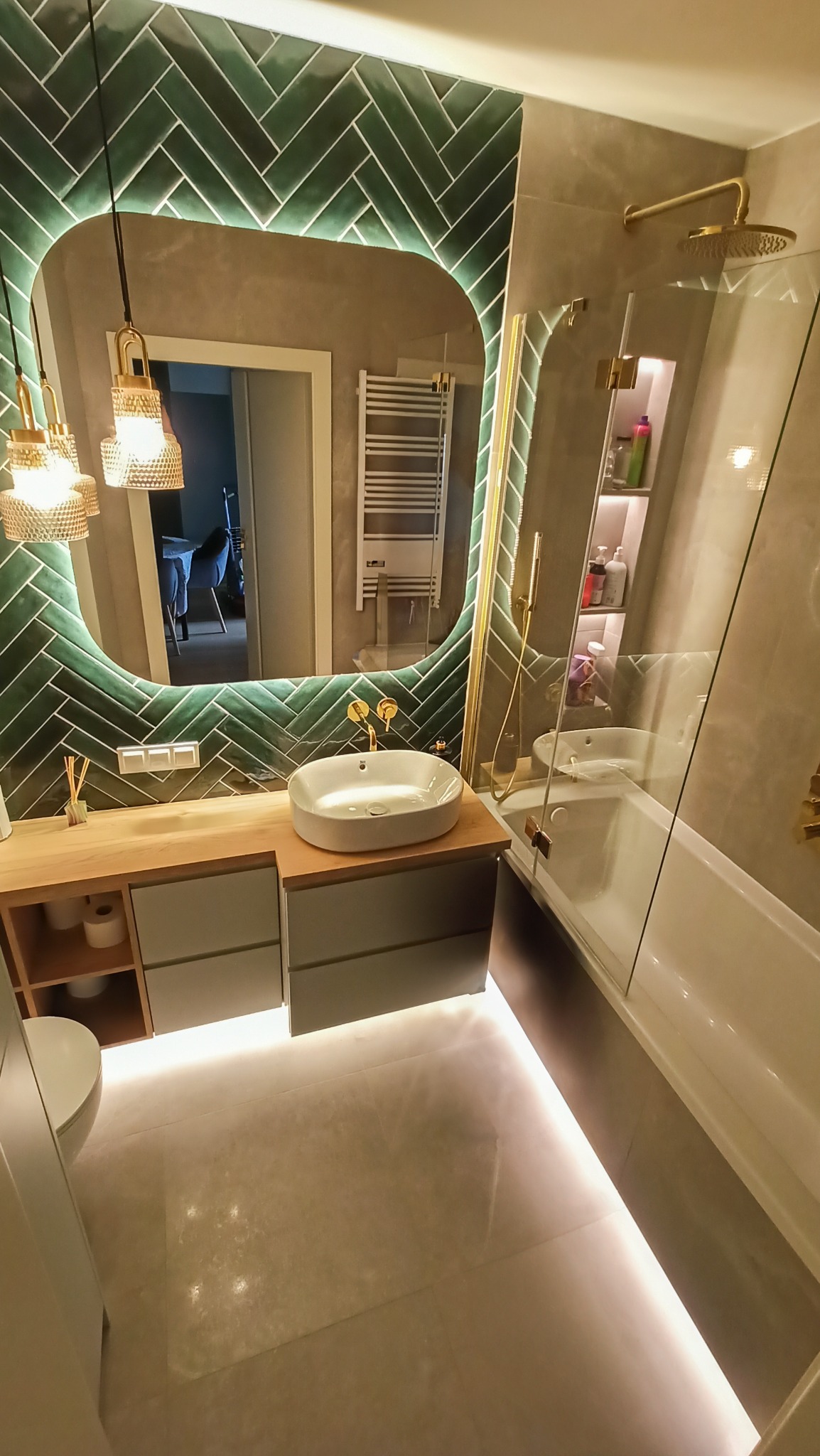 Nowoczesna łazienka z zielonymi płytkami ułożonymi w jodełkę, podświetlanym lustrem i drewnianą szafką z umywalką. Kabina prysznicowa ze złotymi elementami.