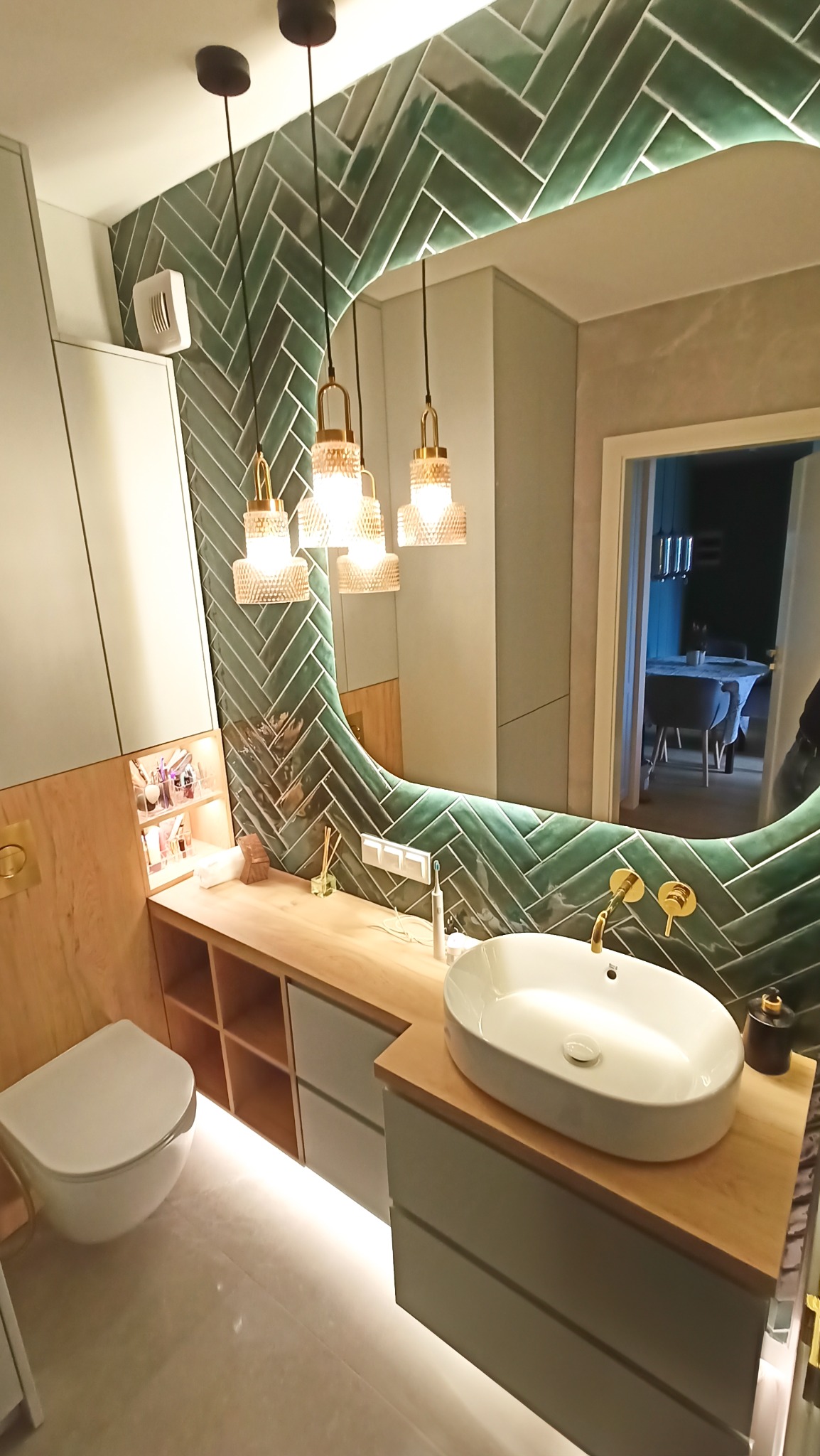 Nowoczesna łazienka z zielonymi płytkami ułożonymi w jodełkę, owalnym lustrem z podświetleniem LED i wiszącą umywalką. Widoczne oświetlenie punktowe i drewniane elementy wykończenia.