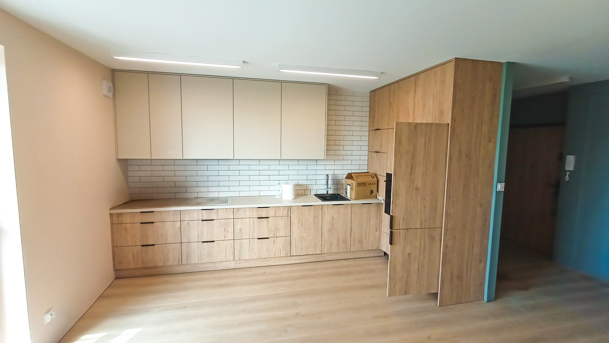 Jasna kuchnia z drewnianymi szafkami i białymi płytkami na ścianie. Widoczny zlew, blat i oświetlenie sufitowe. Minimalistyczny design, świeżo po remoncie.