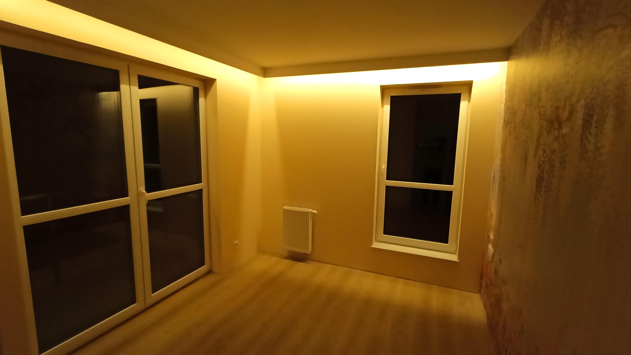 Pokój po remoncie z oknem, drzwiami balkonowymi i grzejnikiem. Ściany pomalowane na beżowo, jedna ściana z efektem dekoracyjnym. Oświetlenie sufitowe.