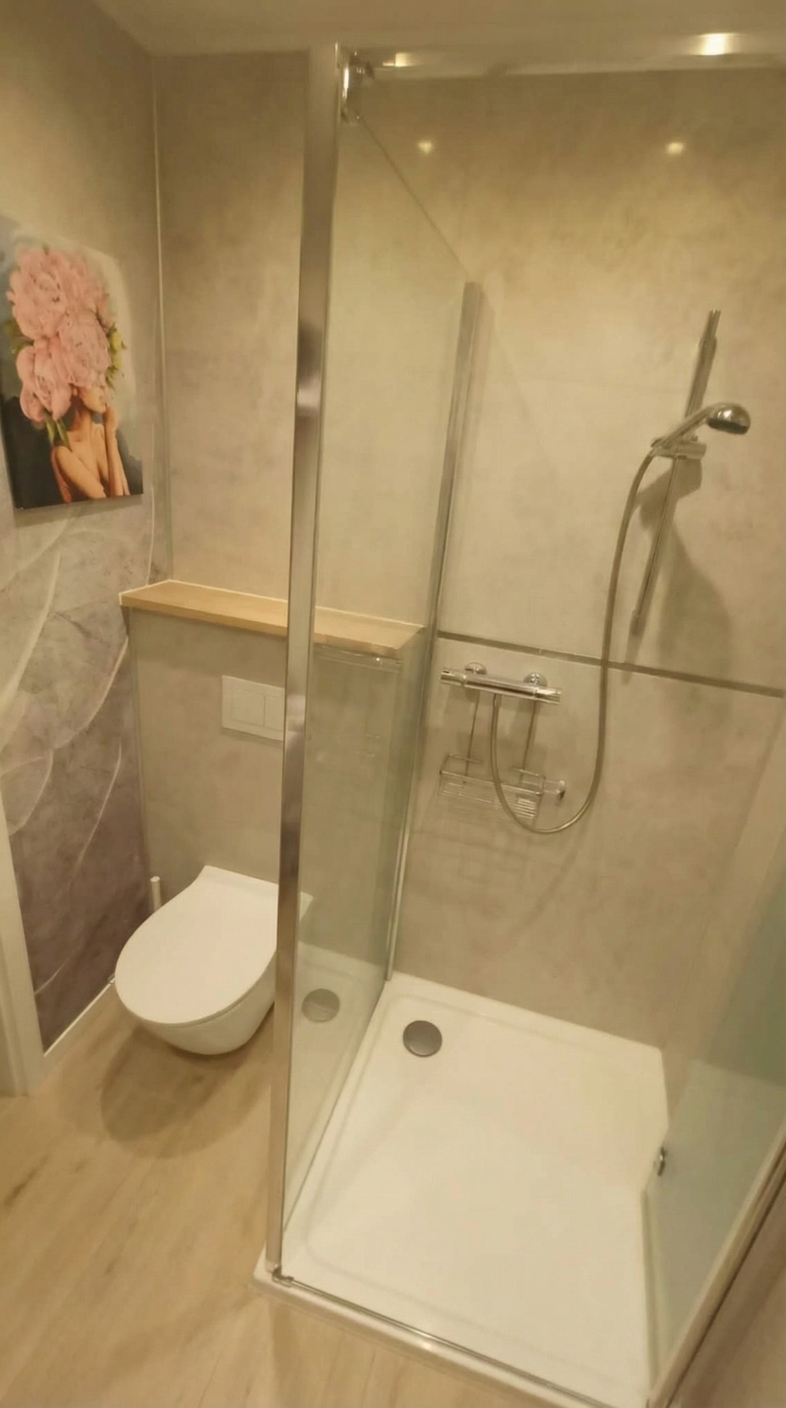 Nowoczesna łazienka z kabiną prysznicową typu walk-in, toaletą podwieszaną i półką z jasnego drewna. Na ścianie grafika z motywem kwiatowym.