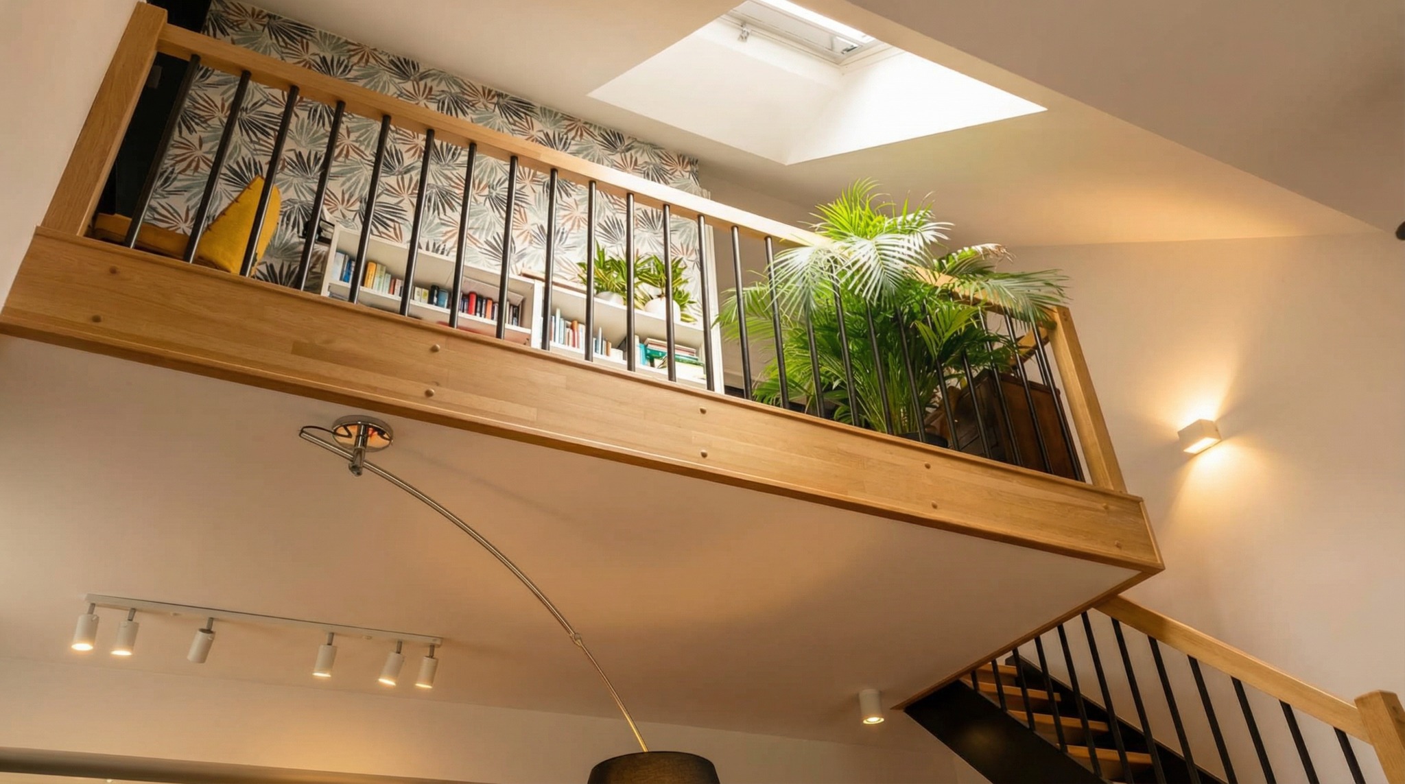 Drewniana antresola z czarnymi balustradami, oświetlona światłem z okna dachowego i lamp, z roślinami i regałem z książkami w tle, widok z dołu.