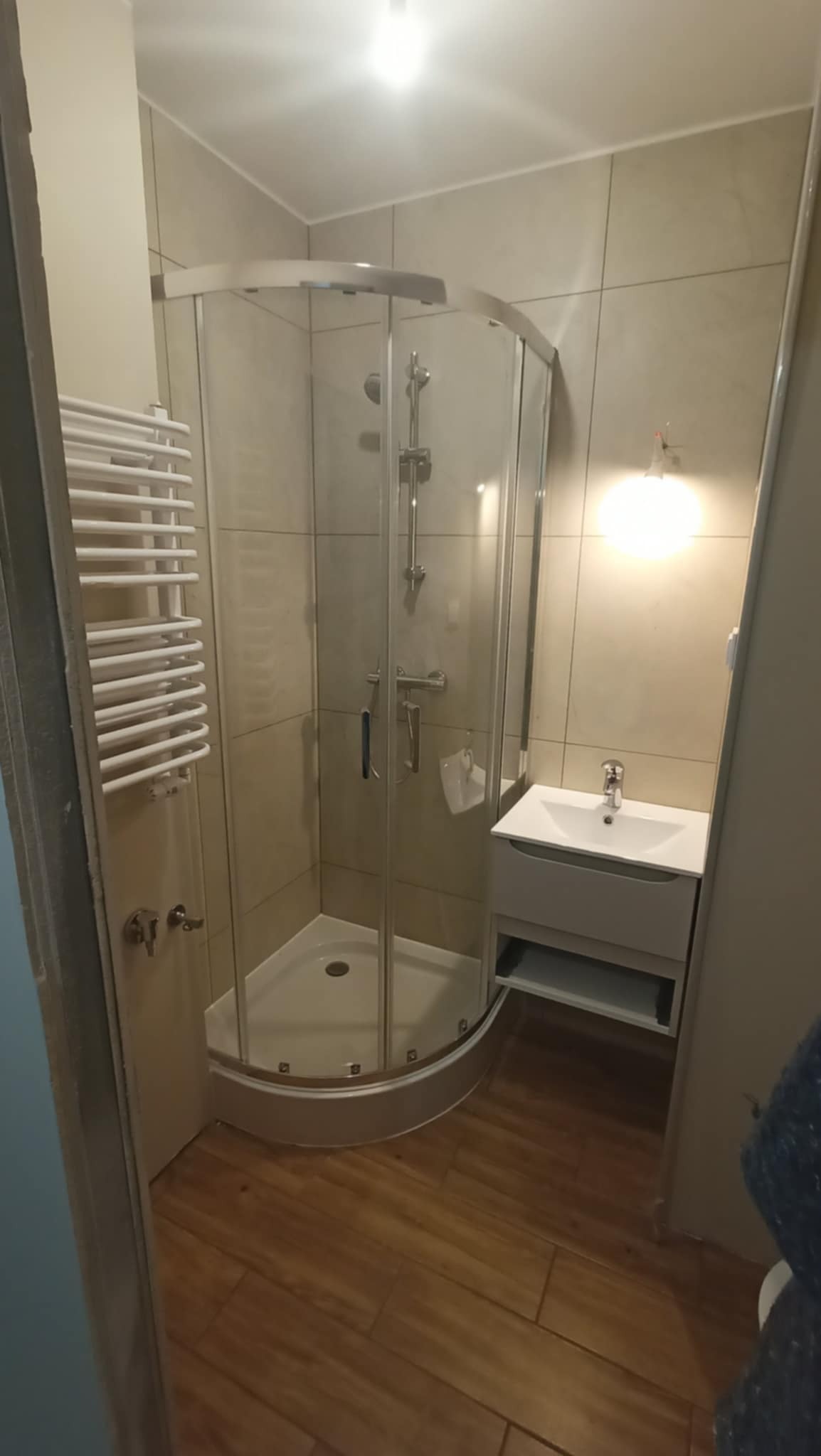 Nowoczesna łazienka po remoncie: kabina prysznicowa narożna, umywalka z szafką, grzejnik drabinkowy, płytki w odcieniach beżu i drewnopodobna podłoga.