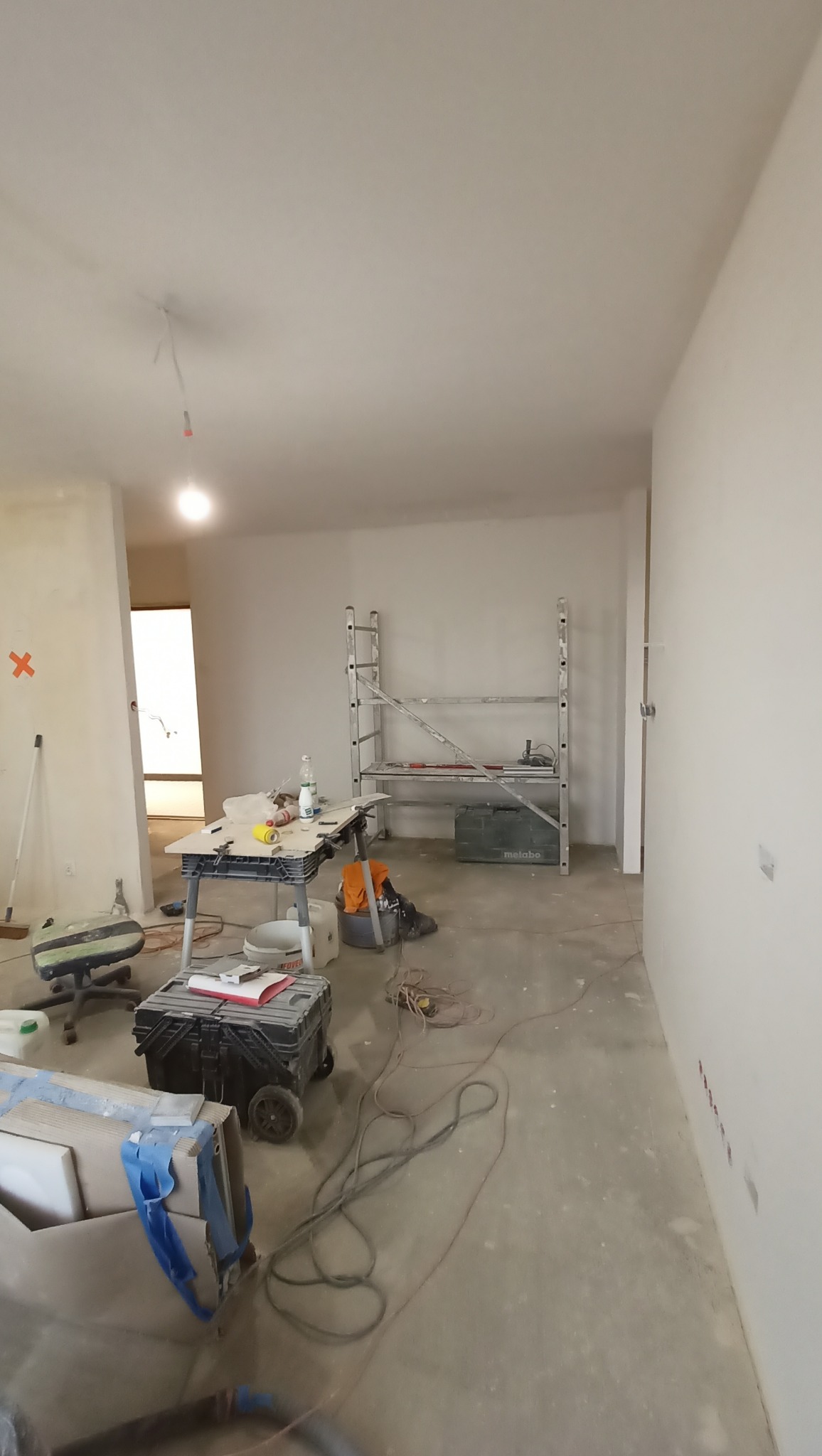 Wnętrze w trakcie remontu: narzędzia, drabina, kable na podłodze. Ściany przygotowane do malowania, widoczne ślady prac wykończeniowych. Jasne, naturalne światło.