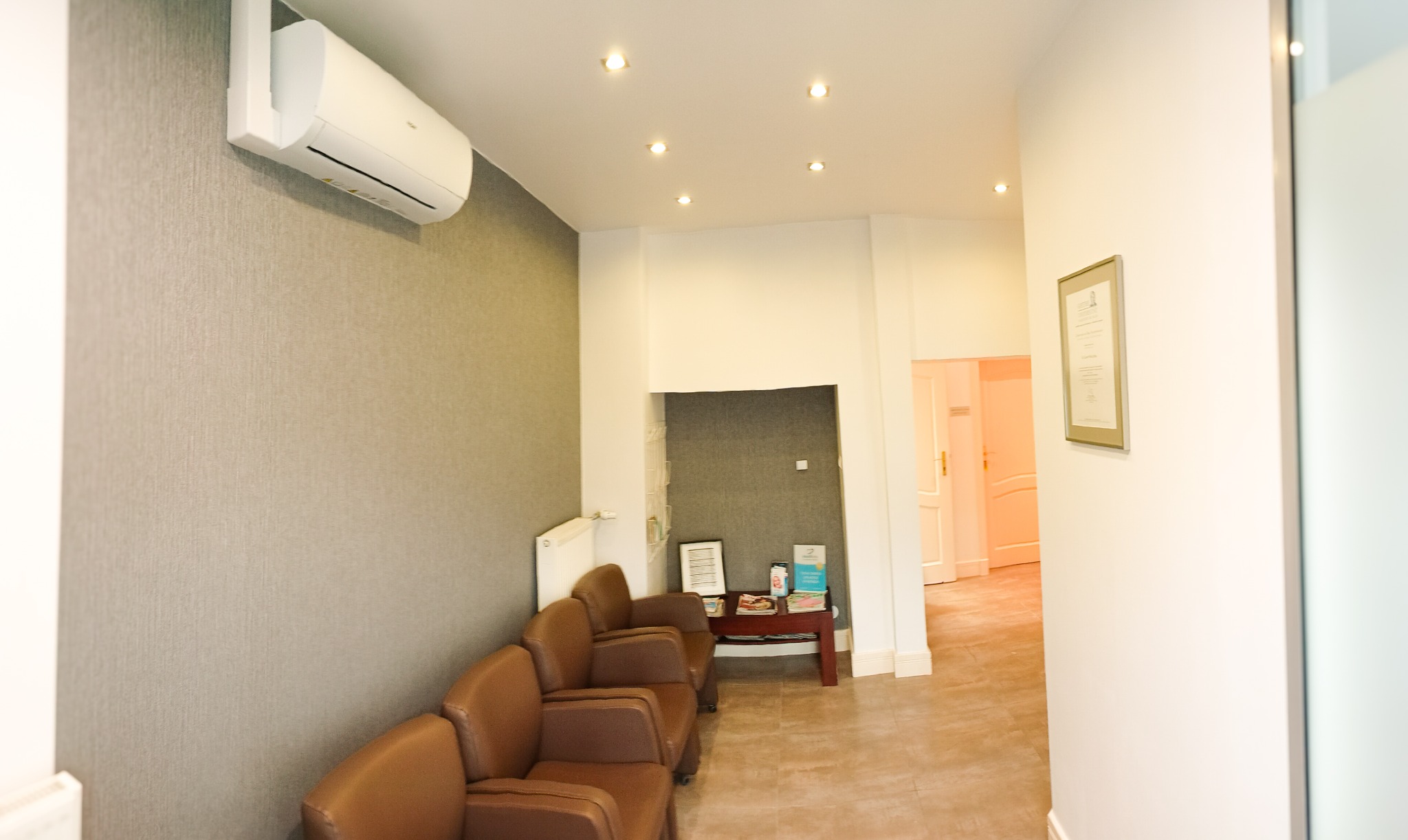 Klimatyzacja na ścianie z tapetą w odcieniach szarości, brązowe fotele w poczekalni, oświetlenie punktowe i certyfikat w ramce na ścianie w tle.