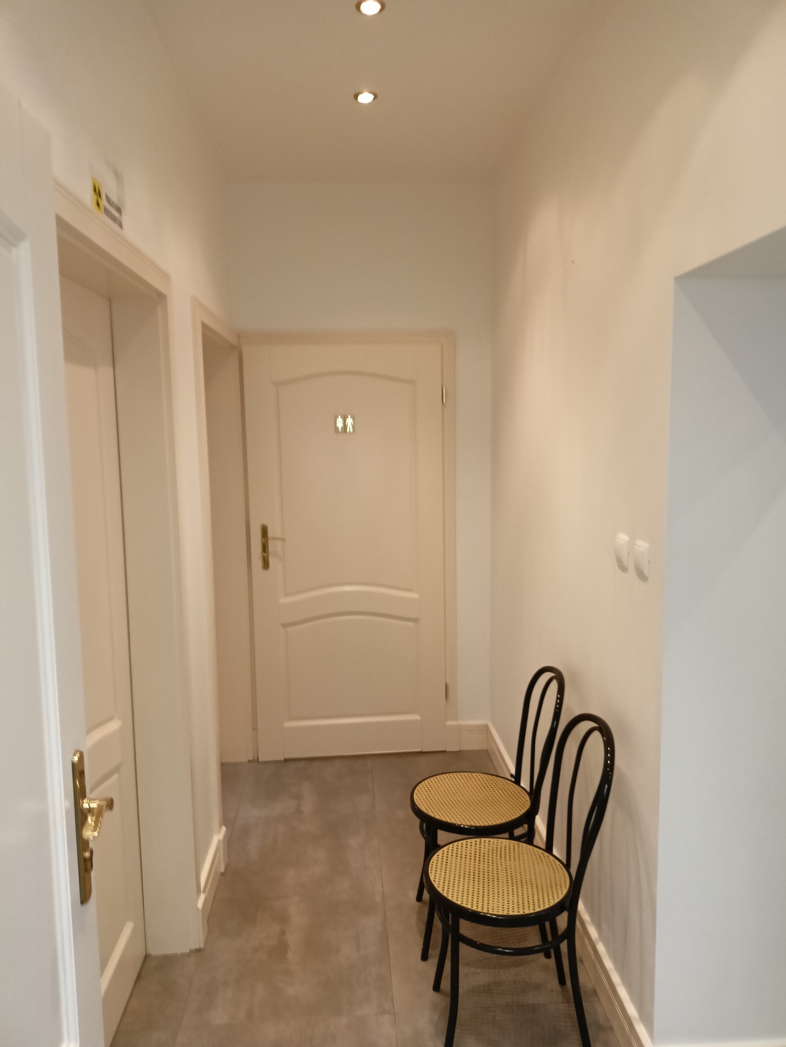 Jasny korytarz z dwiema krzesłami wiedeńskimi, prowadzący do drzwi z oznaczeniem toalet. Minimalistyczny design, szara podłoga, subtelne oświetlenie.