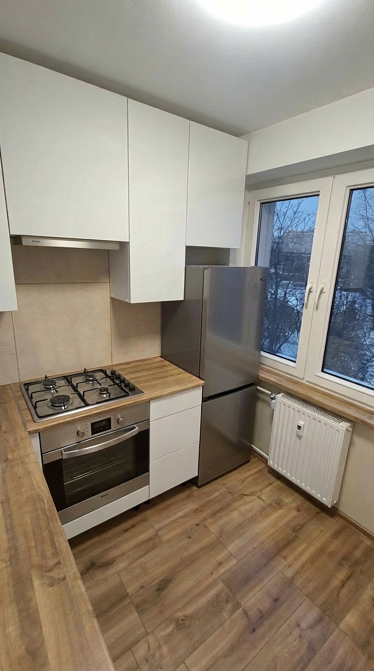 Jasna kuchnia z drewnianą podłogą i białymi szafkami. Widoczna kuchenka, piekarnik i lodówka. Okno z widokiem na drzewa. Minimalistyczny design.