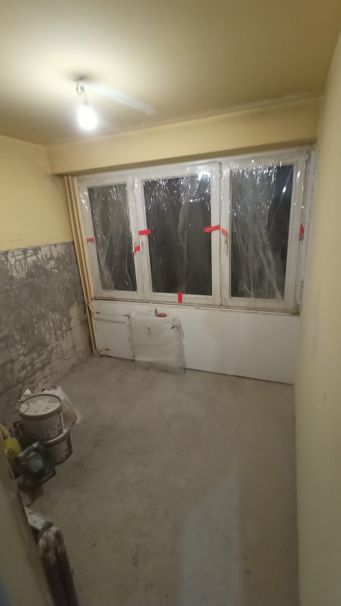 Pokój w trakcie remontu: ściany przygotowane do malowania, okna zabezpieczone folią, widoczne wiadra z farbą. Przygotowanie do prac wykończeniowych.