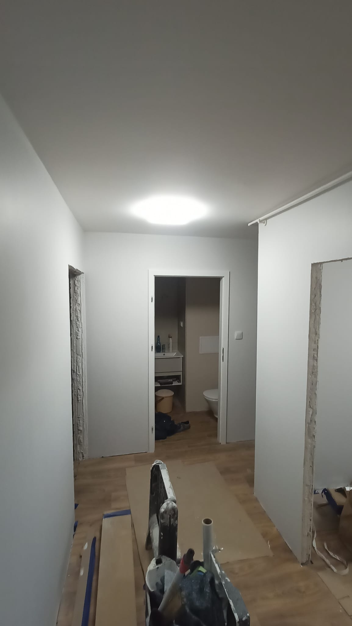 Wnętrze w trakcie remontu: świeżo pomalowane ściany na szaro, widoczne narzędzia malarskie, przygotowane wejście do łazienki. Podłoga zabezpieczona kartonem.
