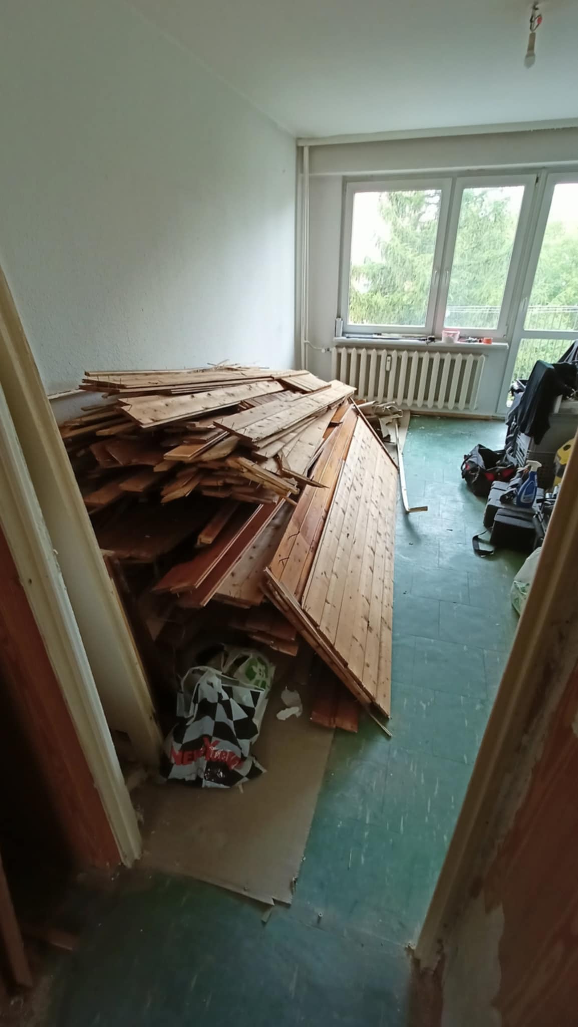 Stos drewnianych paneli podłogowych w pokoju przed remontem. Widoczne okno, grzejnik i narzędzia. Podłoga w odcieniach zieleni. Przygotowanie do prac remontowych.