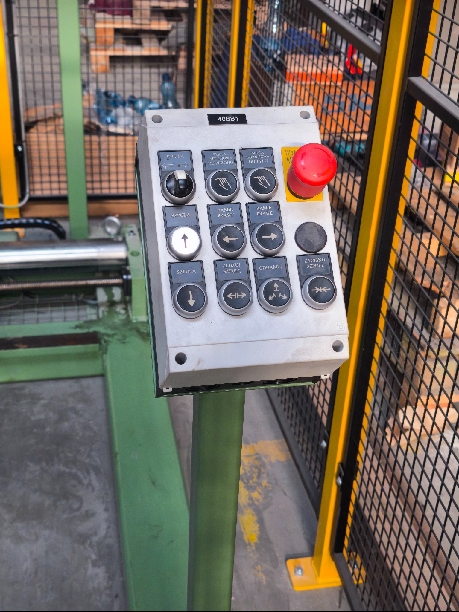Panel sterowania maszyny przemysłowej z przyciskami funkcyjnymi i wyłącznikiem awaryjnym, umieszczony na zielonym słupku, z metalową siatką w tle.