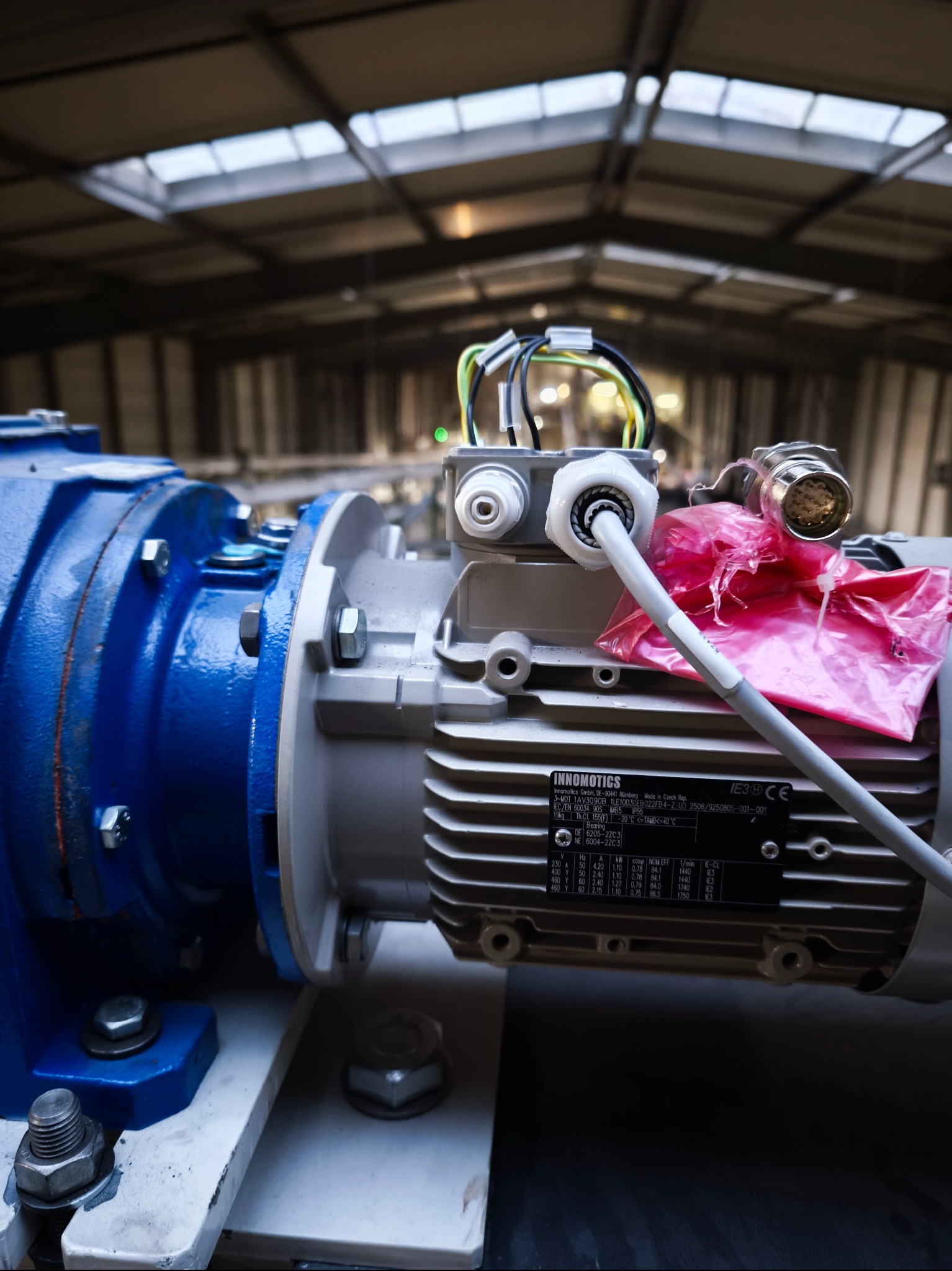 Szczegółowe ujęcie silnika elektrycznego Innomotics z podłączonymi kablami, umieszczonego w hali przemysłowej z oknami dachowymi. Widoczne śruby i elementy mocujące.