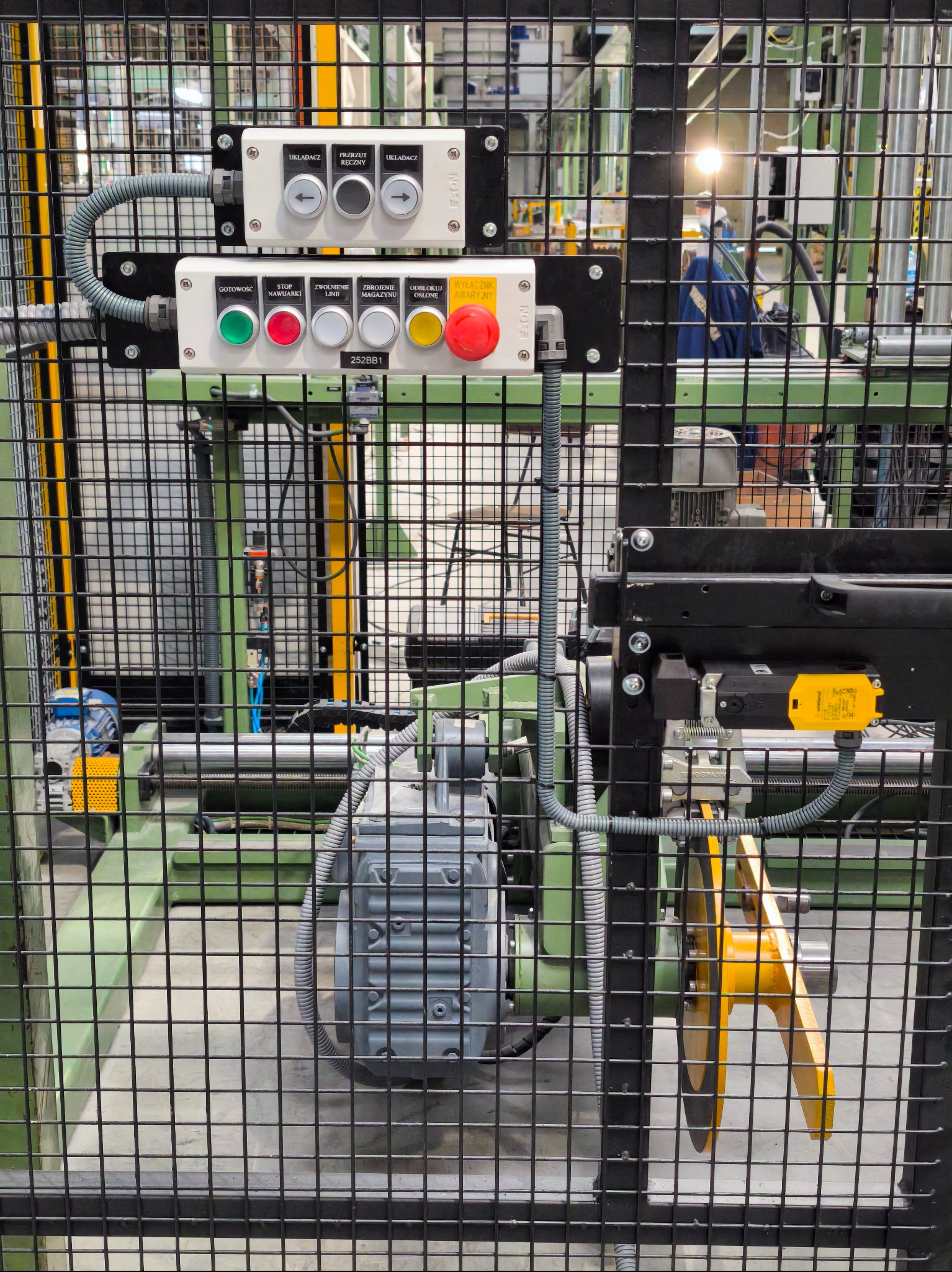 Panel sterowania maszyny przemysłowej za metalową siatką ochronną. Widoczne przyciski, przełączniki i okablowanie. Szara obudowa silnika w dolnej części kadru.