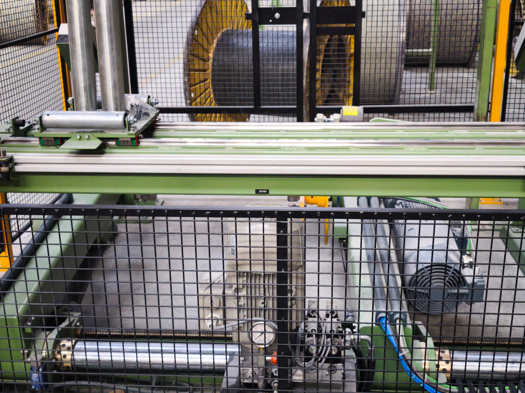 Zabezpieczona siatką maszyna do produkcji kabli z widocznymi bębnami z nawiniętym kablem, silnikiem i elementami sterującymi. Detal z bliska.