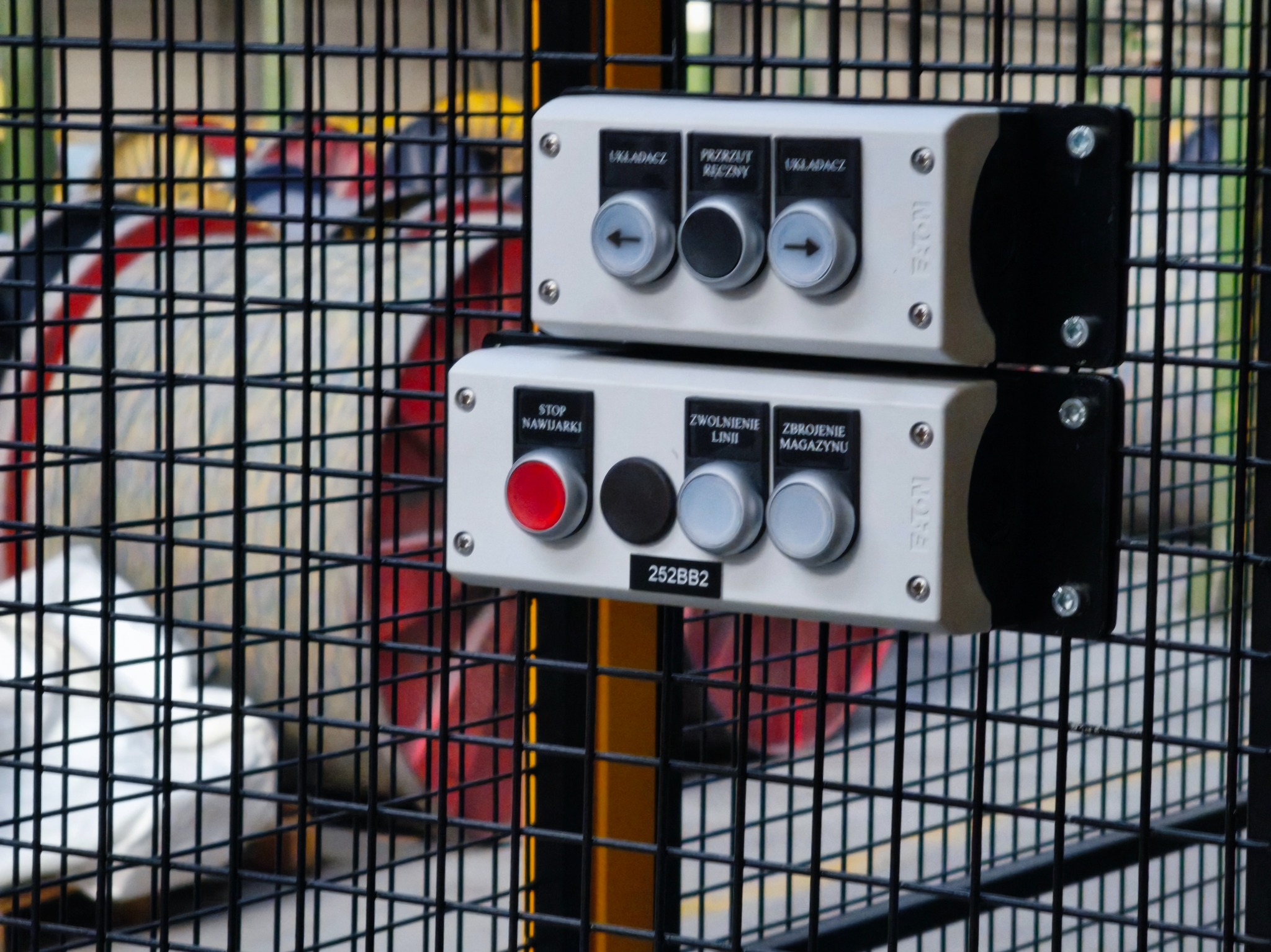 Panel sterowania maszyny z przyciskami i pokrętłami, umieszczony za metalową siatką ochronną. Widoczne napisy: 'Stop', 'Zwolnienie linii', 'Zbrojenie magazynu'.