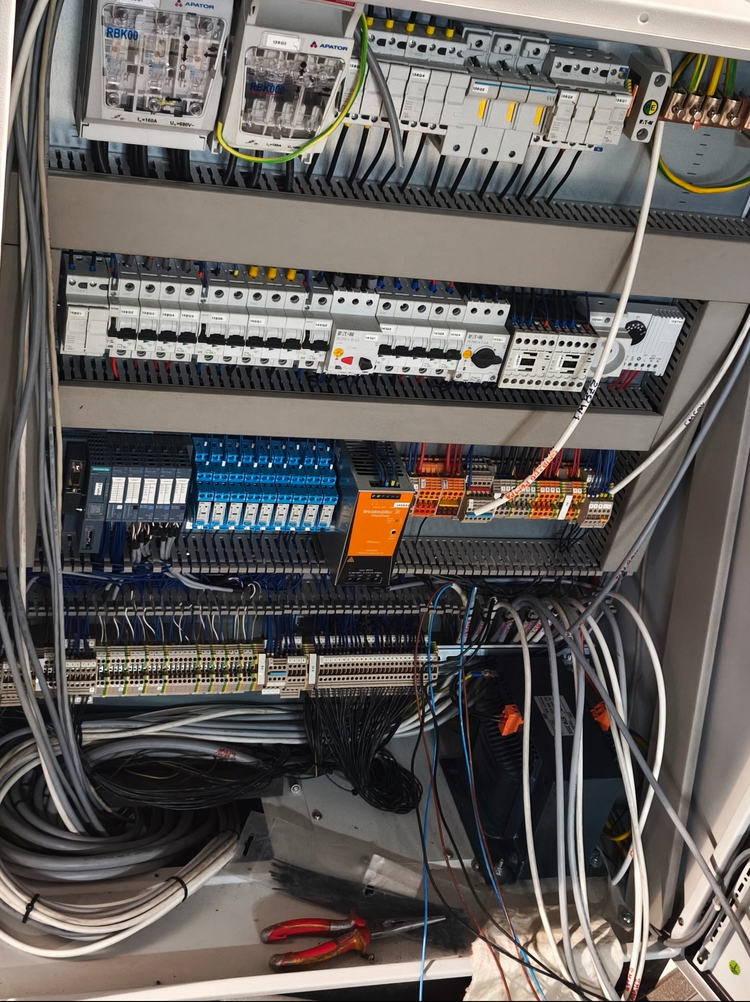 Rozdzielnica elektryczna z widocznymi bezpiecznikami, przekaźnikami i okablowaniem. Widoczne narzędzia. Detal montażu instalacji w szarej obudowie.