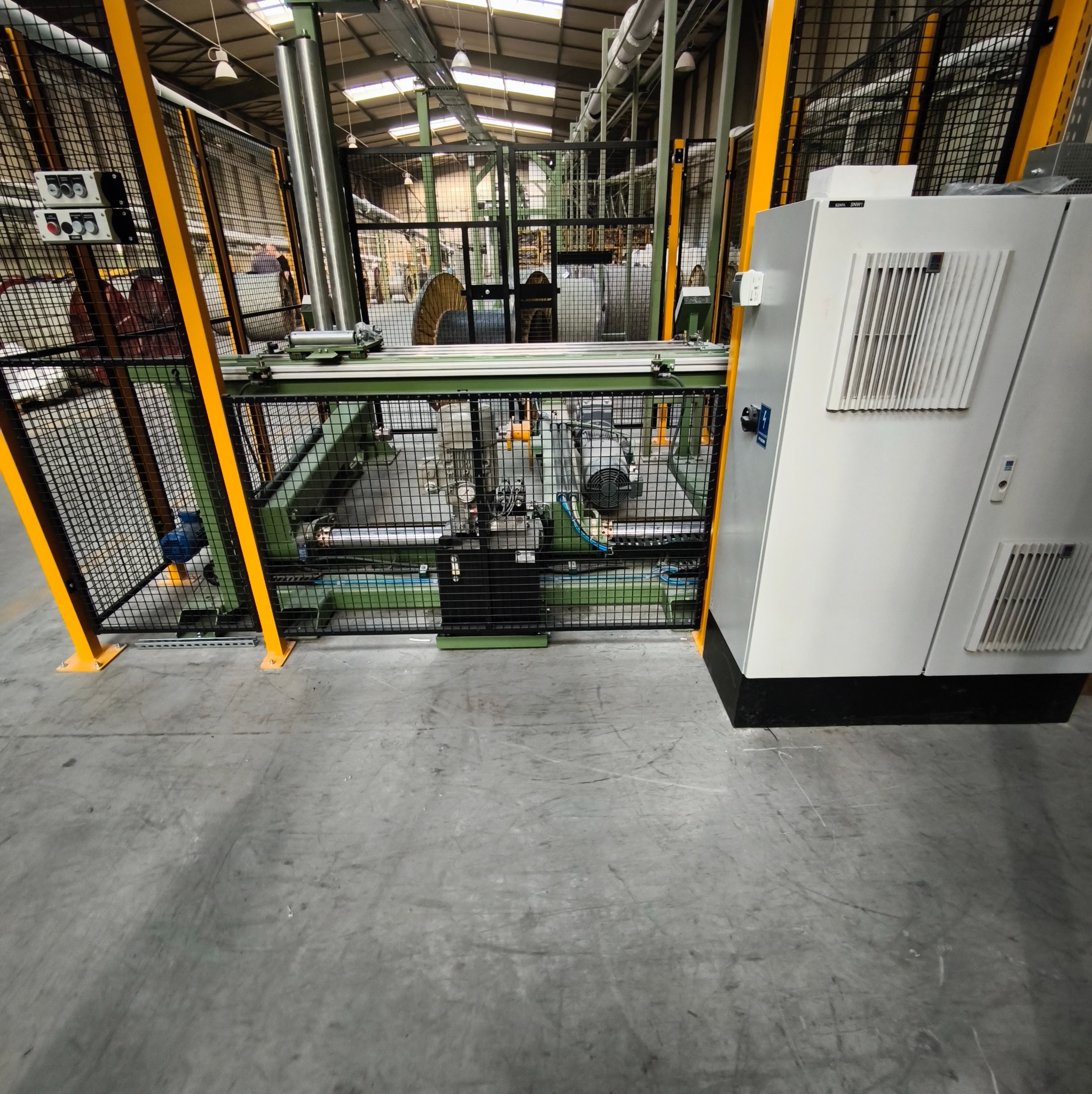 Panel sterowania elektrycznego w hali produkcyjnej, zabezpieczony żółtą metalową siatką. Widoczne kable i elementy instalacji. Szara betonowa podłoga.