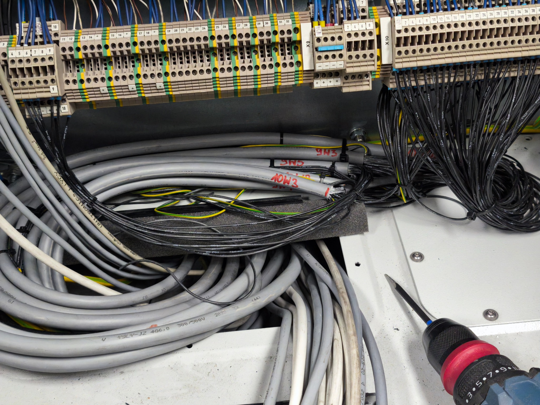 Rozdzielnica elektryczna z licznymi przewodami i złączkami, widoczne kable szare i czarne, oraz wkrętak z czerwonym uchwytem. Instalacja w trakcie prac.