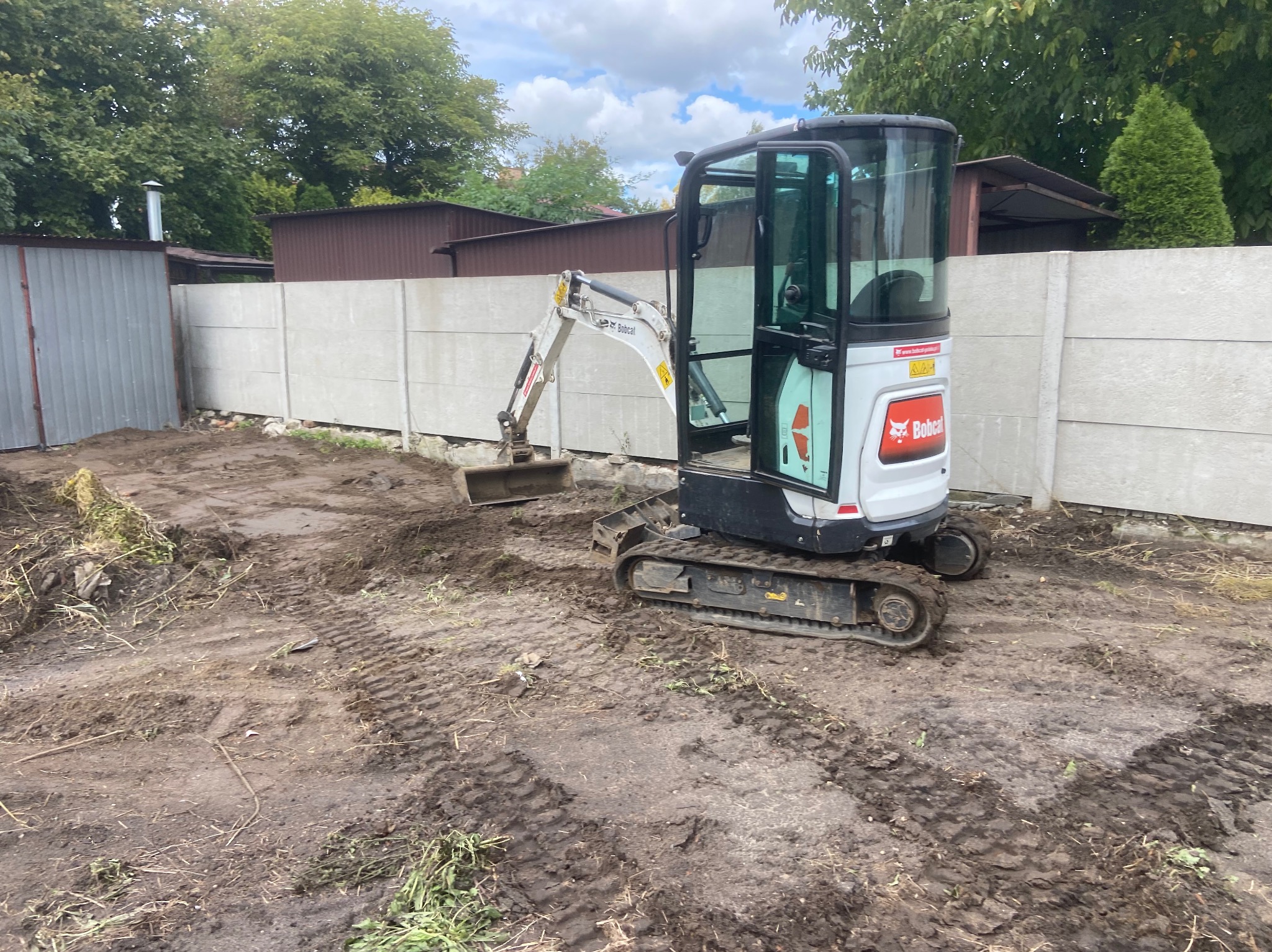 Mini koparka Bobcat na placu budowy, ślady opon w błocie, betonowe ogrodzenie w tle, częściowo oczyszczony teren. Prace ziemne w toku.