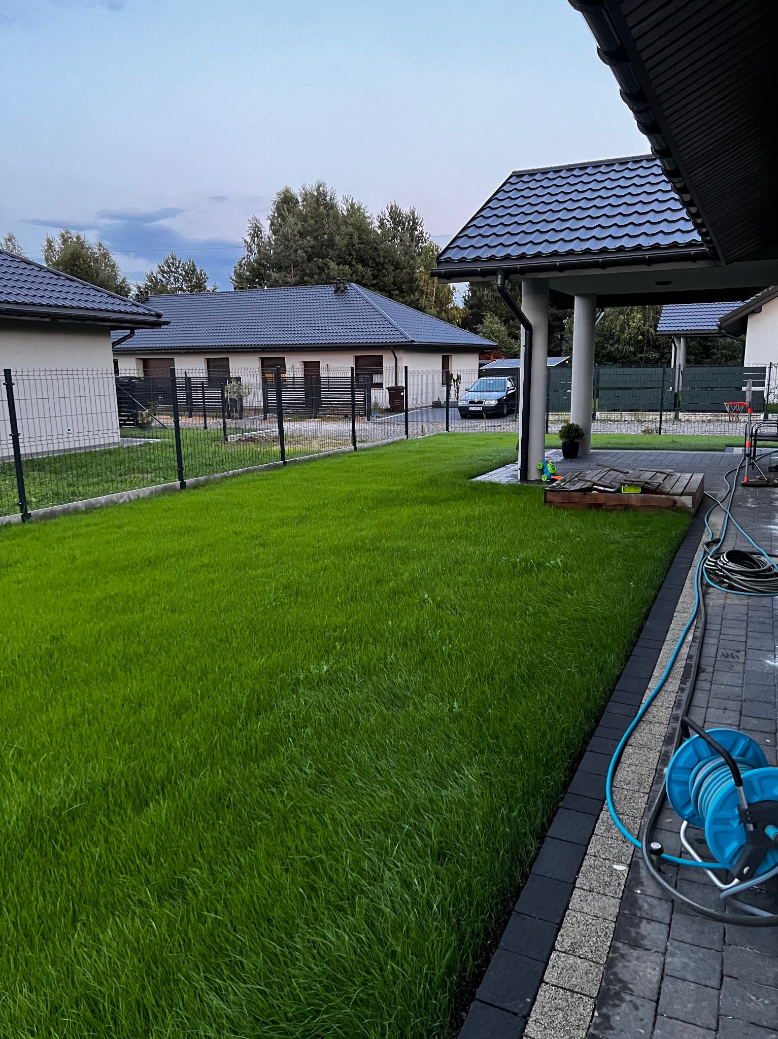Soczysta, zielona trawa w ogrodzie z nowoczesnym domem w tle, widok z boku, z elementami nawadniania na pierwszym planie. Świeżo założony trawnik.