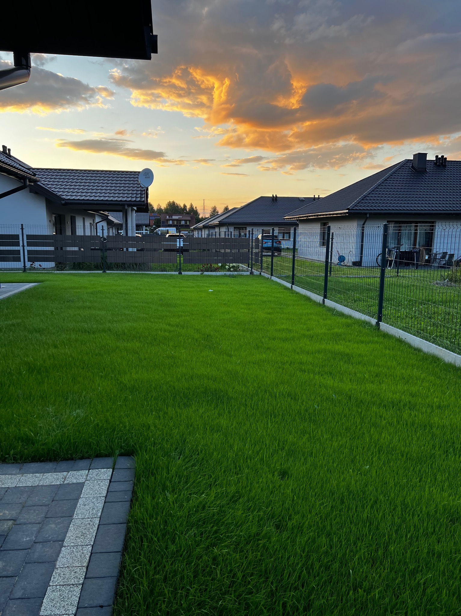Zadbany trawnik przed domem jednorodzinnym, widok z poziomu kostki brukowej, w tle zachodzące słońce barwi chmury na pomarańczowo.