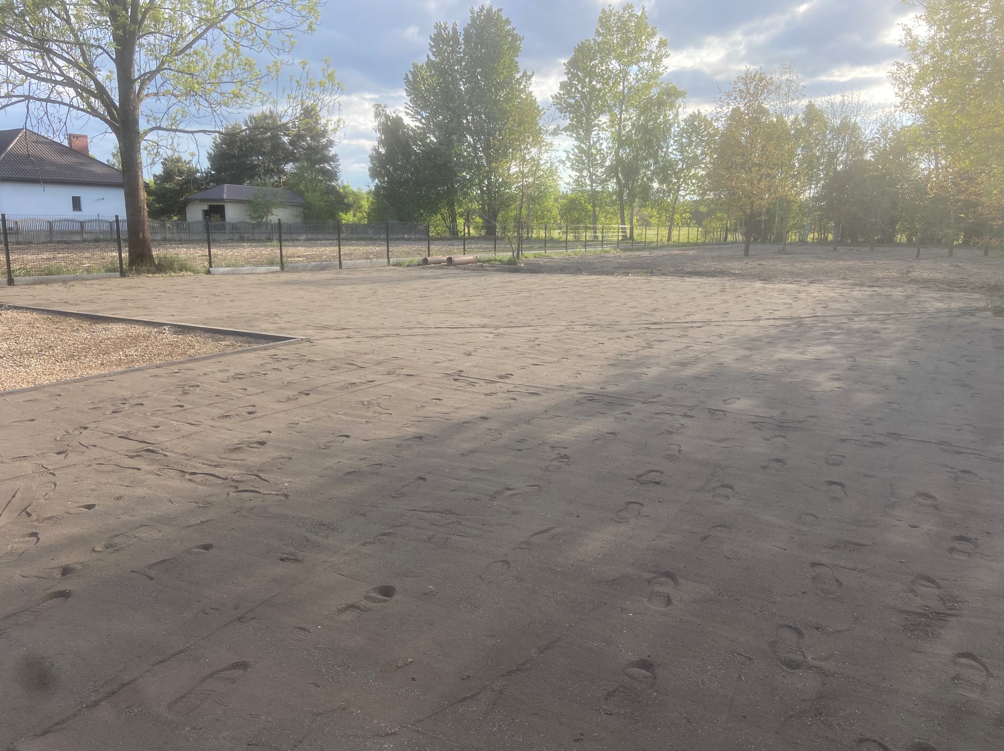 Przygotowany teren pod trawnik z widocznymi śladami stóp, obok budynek mieszkalny i drzewa. Utwardzona droga z kostki po lewej stronie.