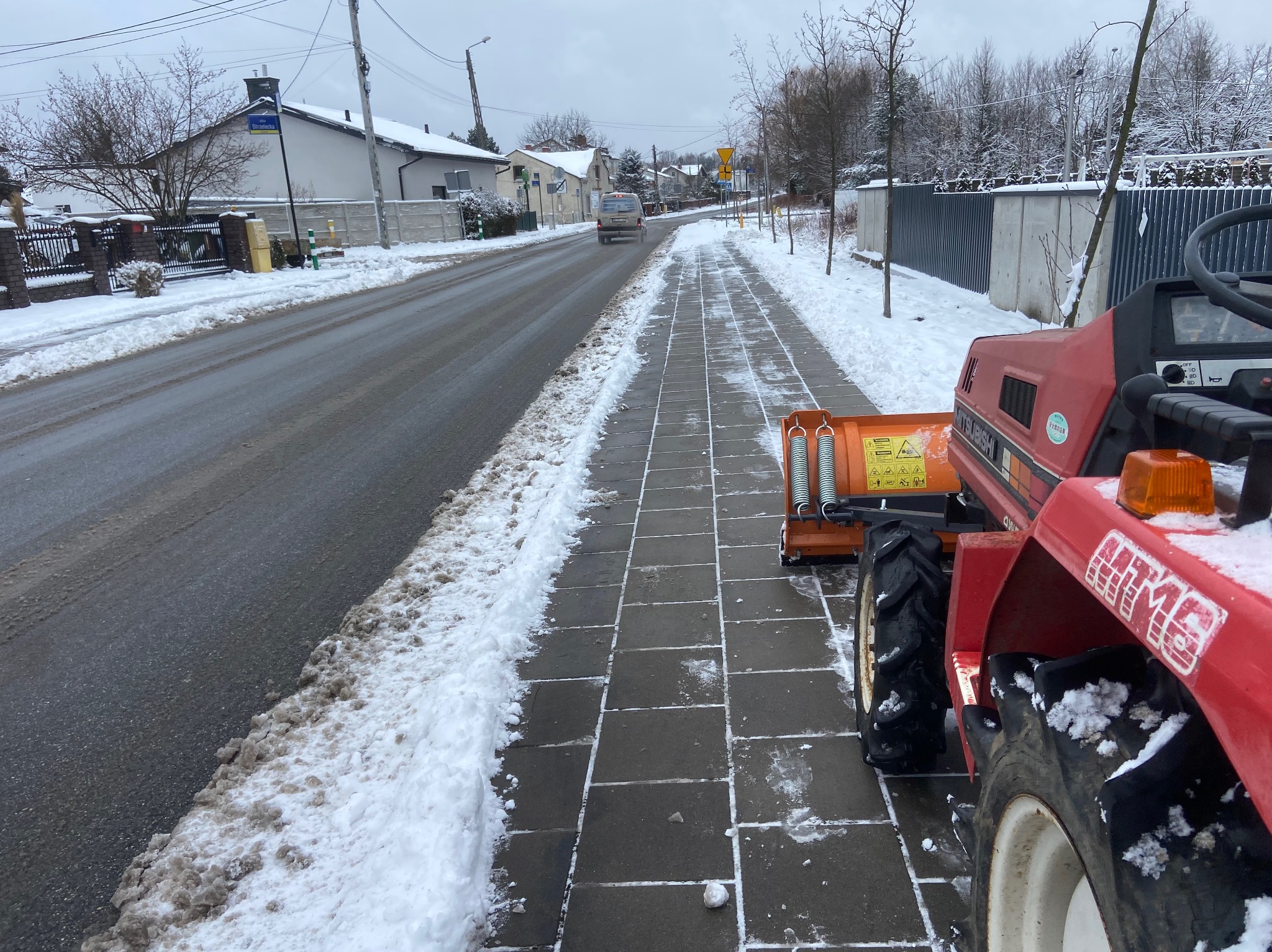 Czerwony traktor z pługiem odśnieża chodnik z kostki brukowej w zimowy dzień. Na jezdni widać resztki śniegu, w tle zabudowa jednorodzinna i pochmurne niebo.
