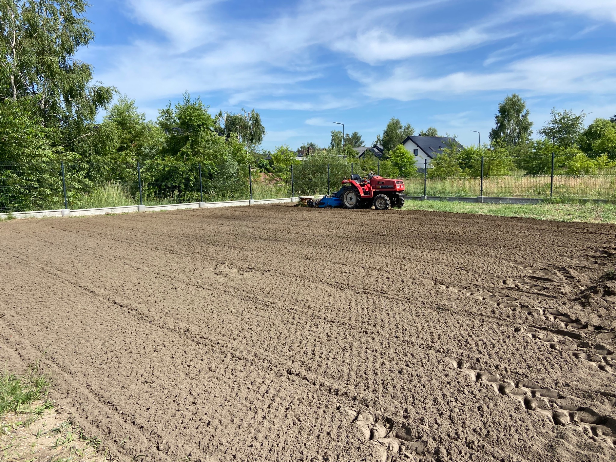 Czerwony traktor z glebogryzarką przygotowuje ziemię pod zasiew trawy na polu. W tle drzewa i domy.