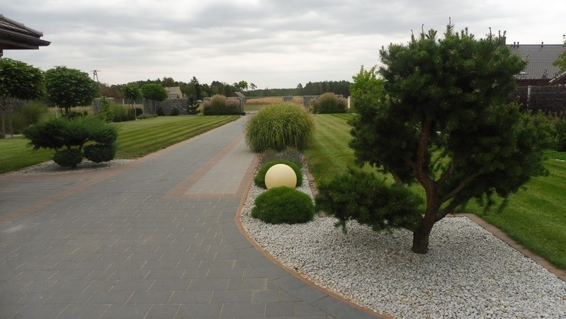 Uporządkowany ogród z równo przystrzyżonym trawnikiem, ozdobnymi krzewami i ścieżką z kostki brukowej. Dekoracyjny kamień i kula ogrodowa dodają elegancji.