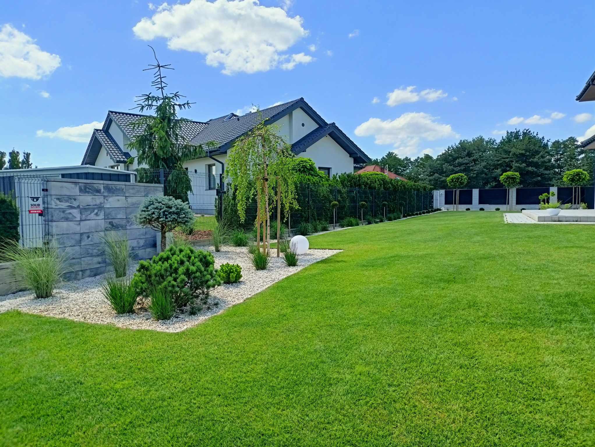 Zadbany ogród z równo przystrzyżonym trawnikiem, ozdobnymi krzewami i drzewami, białymi kamieniami oraz nowoczesnym domem w tle, w słoneczny dzień.