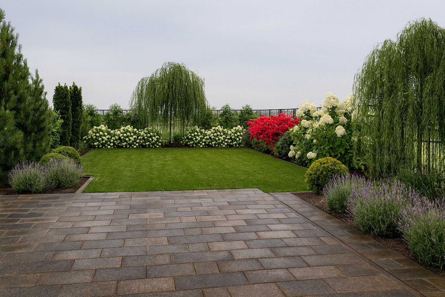 Ułożona kostka brukowa przechodzi w zadbany trawnik, otoczony kwitnącymi krzewami i drzewami, tworząc harmonijny krajobraz ogrodu.