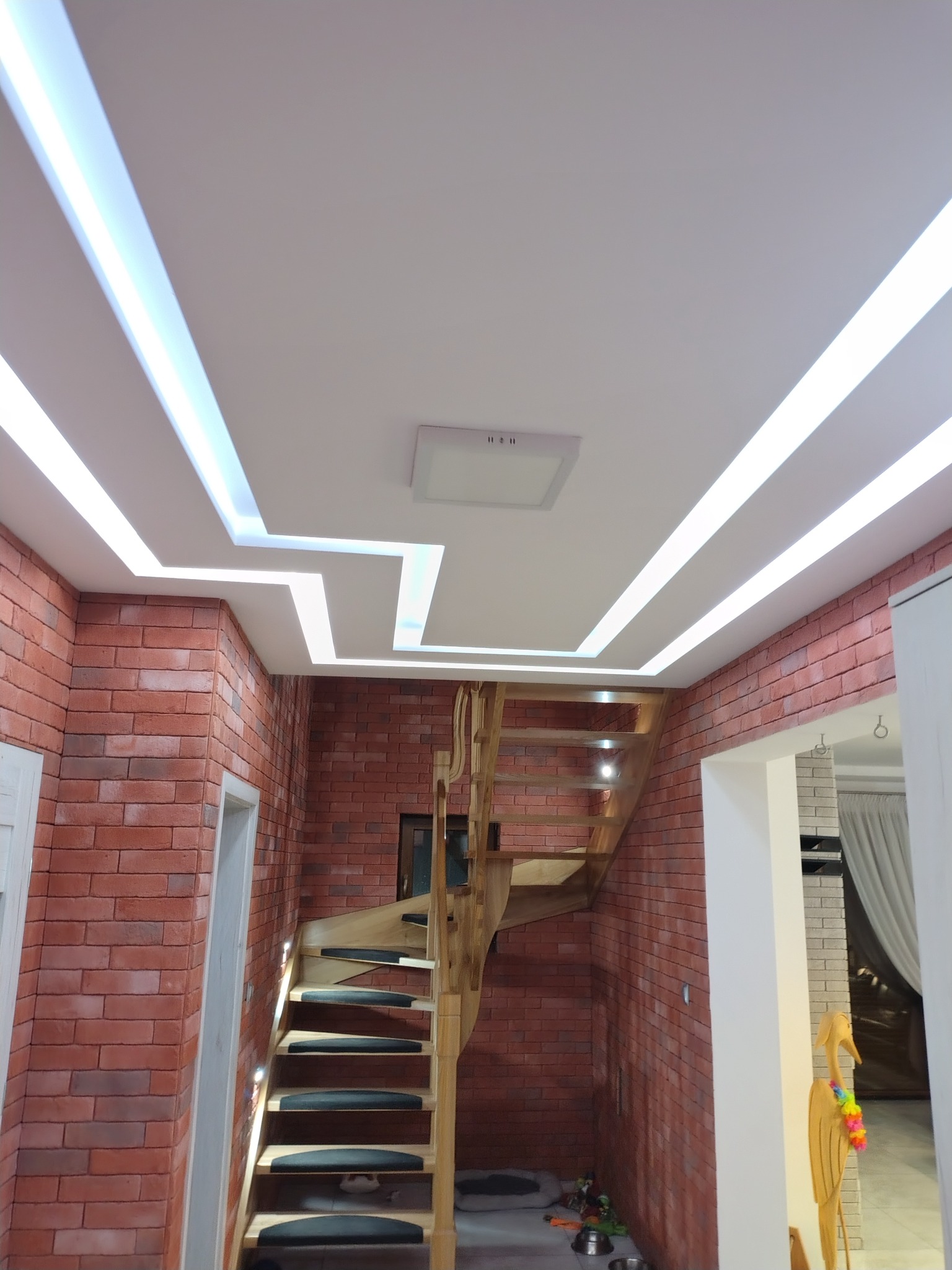 Ceglana ściana z drewnianymi schodami i nowoczesnym oświetleniem sufitowym LED w kształcie zygzaka, tworzącym unikalny design wnętrza.