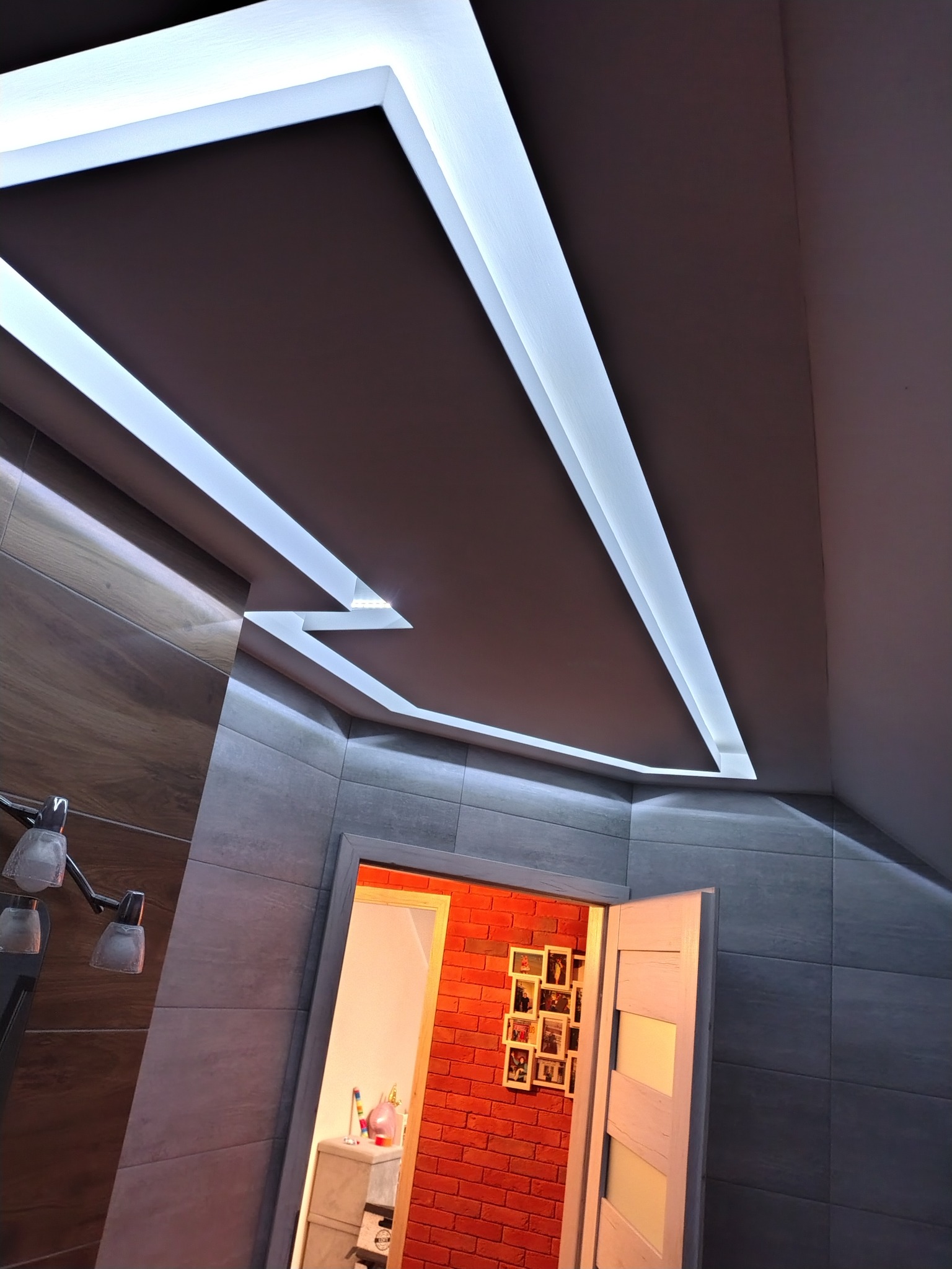 Podwieszany sufit z oświetleniem LED w łazience z szarą glazurą i widokiem na pokój z czerwoną cegłą. Nietypowy kształt sufitu.