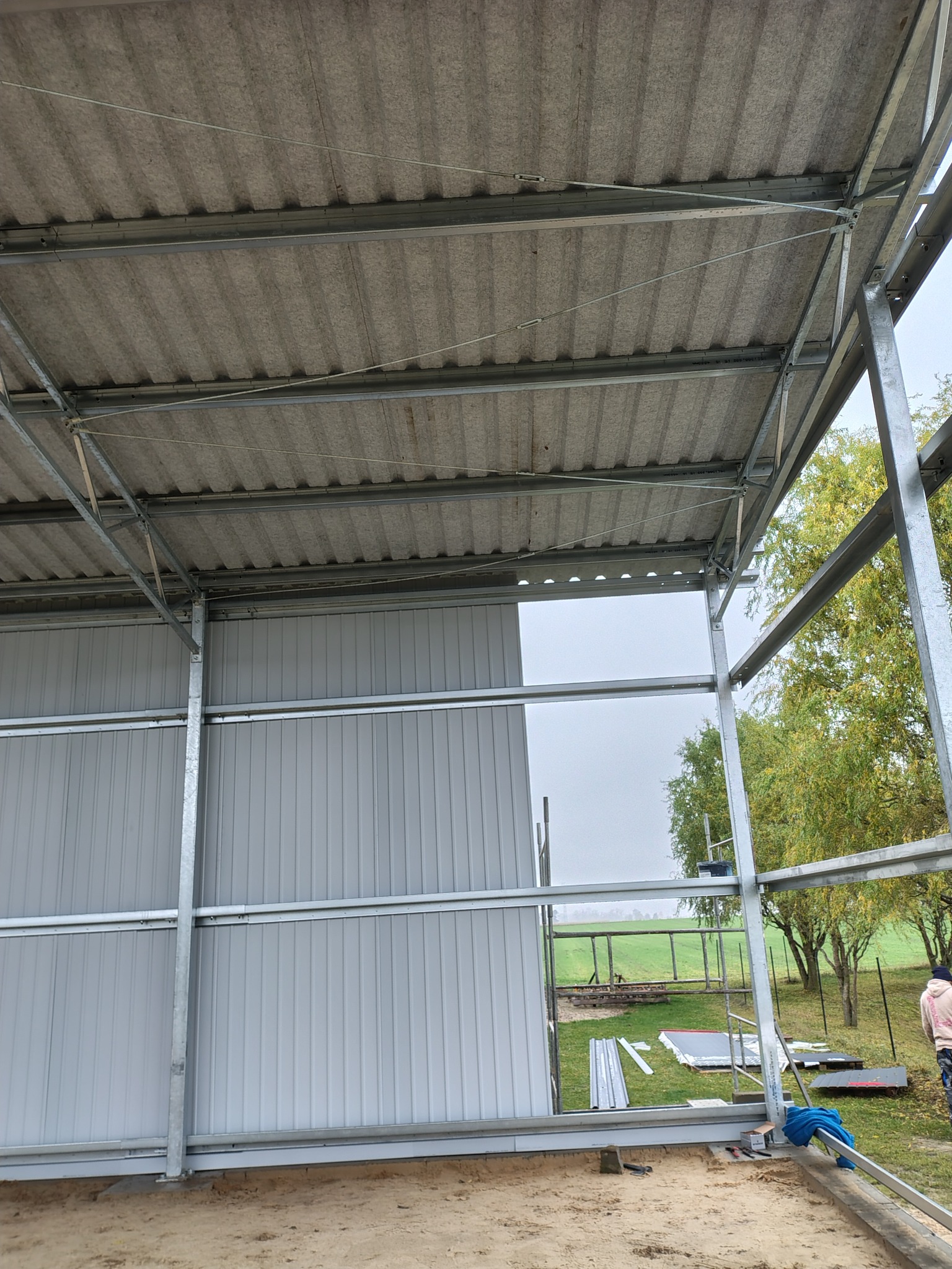 Konstrukcja stalowa z szarą blachą trapezową, widok od wewnątrz. Elementy montażowe, narzędzia i fragment krajobrazu w tle. Budowa w toku, surowy stan.