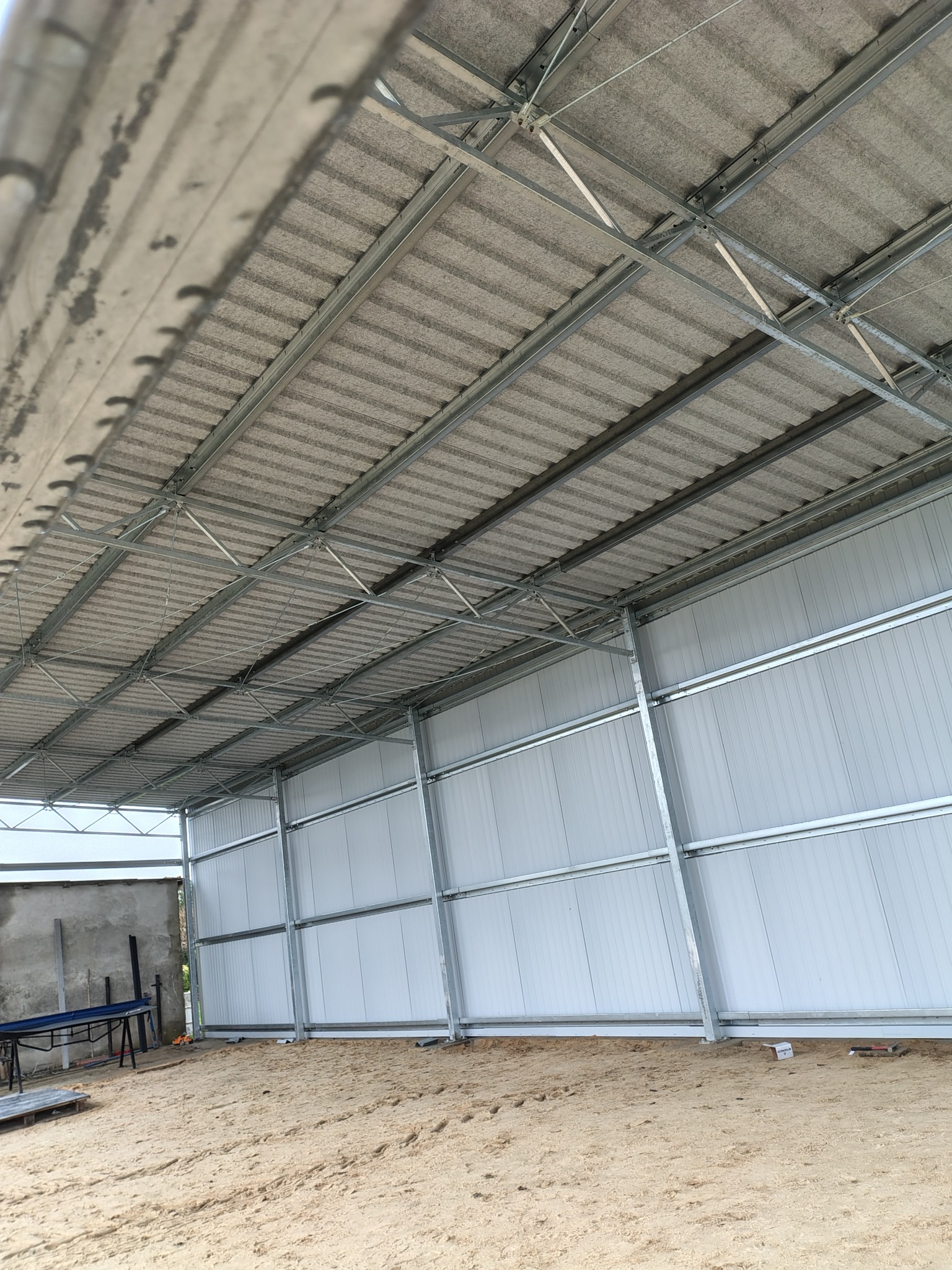 Konstrukcja stalowa hali w trakcie budowy: widoczne ściany z blachy trapezowej, szkielet dachu z metalowych belek i pokrycie dachowe. Piaszczyste podłoże.