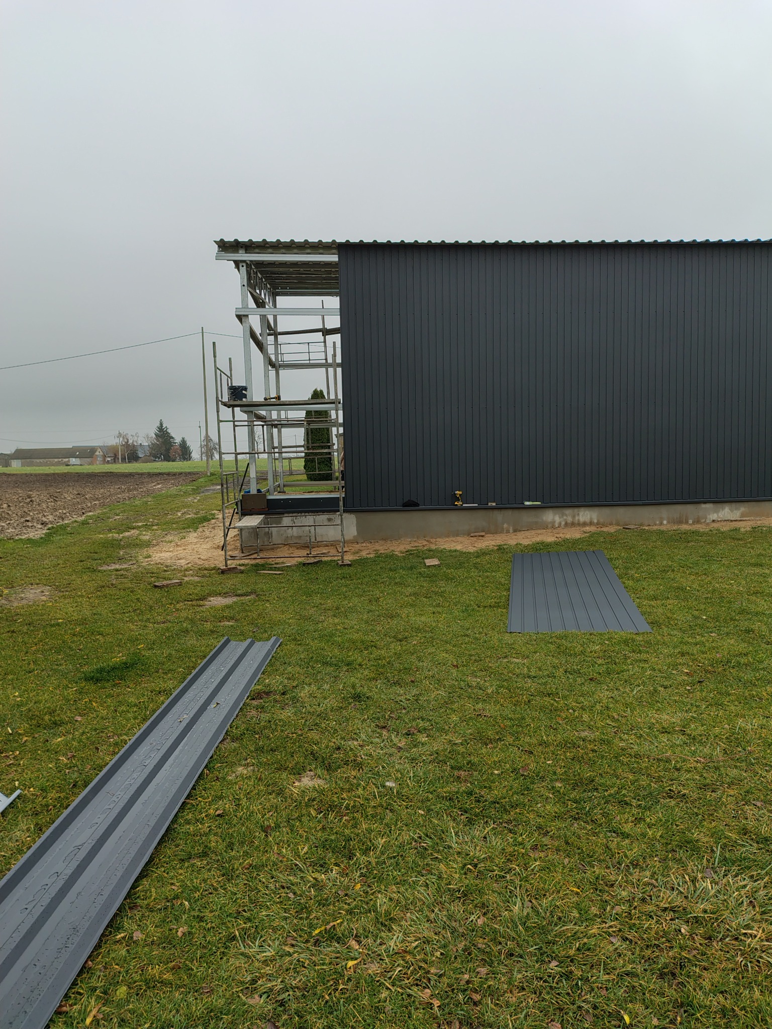 Budowa hali: szare panele ścienne, rusztowanie, narzędzia. Na trawie leżą panele. W tle pole i pochmurne niebo. Widok z poziomu gruntu.