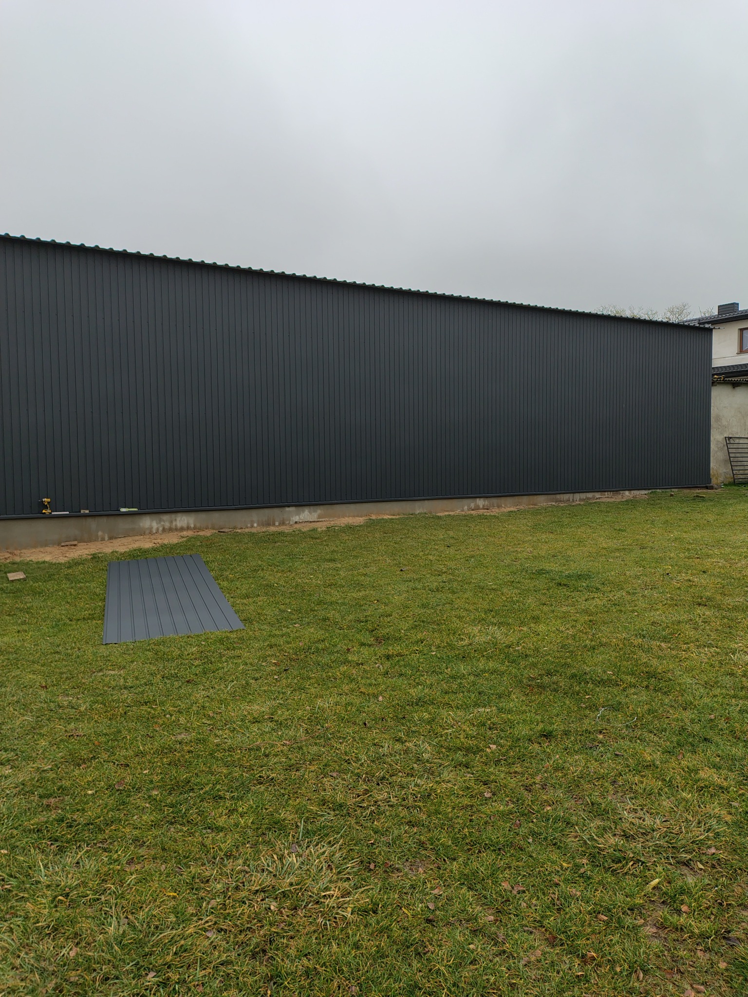 Długa, grafitowa ściana budynku z blachy trapezowej, widoczna z zewnątrz na tle trawnika. Na trawie leży arkusz blachy. W tle fragment budynku sąsiada.