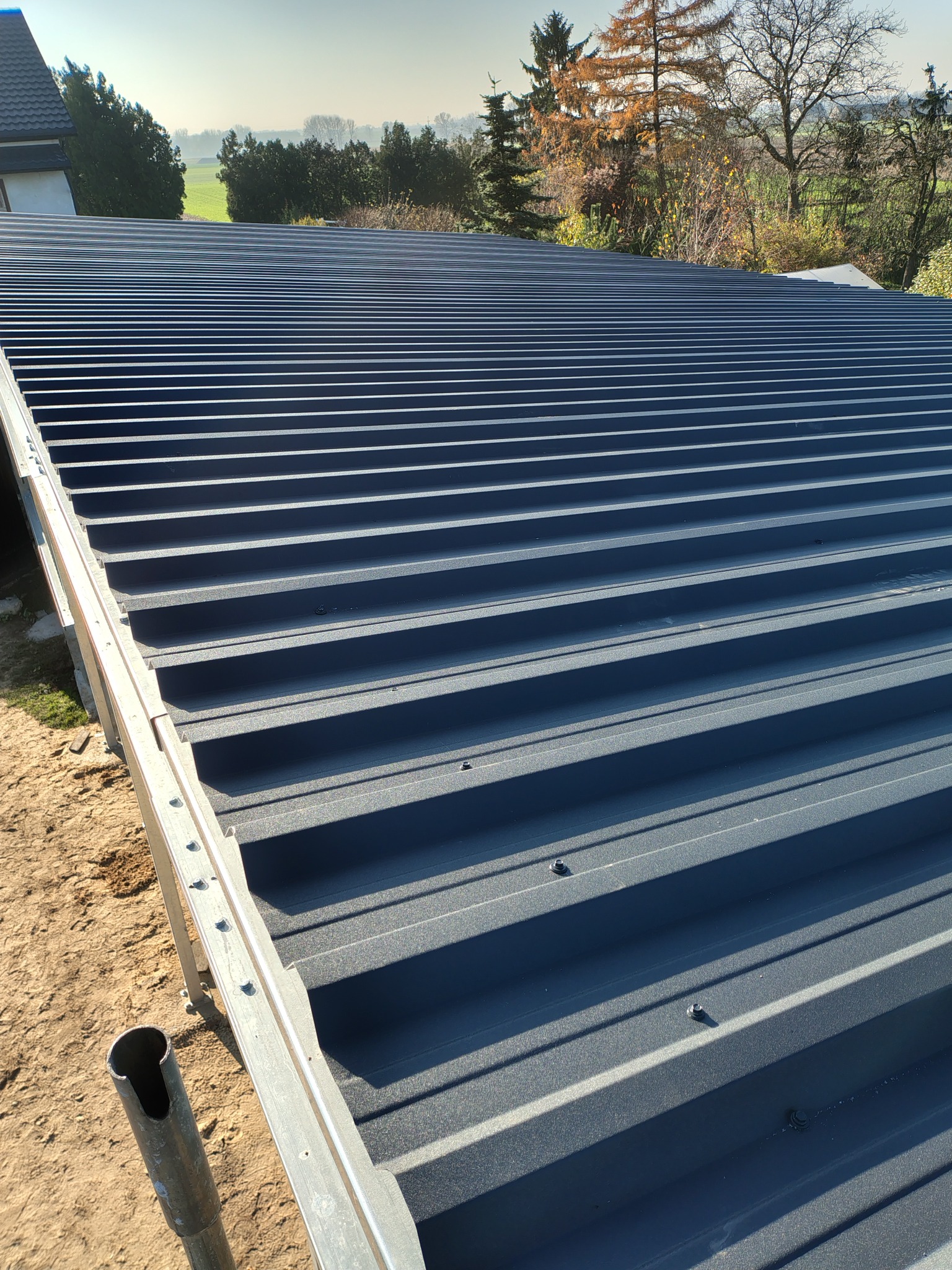 Nowe, grafitowe krycie dachu z blachy trapezowej, widoczne mocowania. W tle drzewa i błękitne niebo. Widok z góry na ukończony dach.