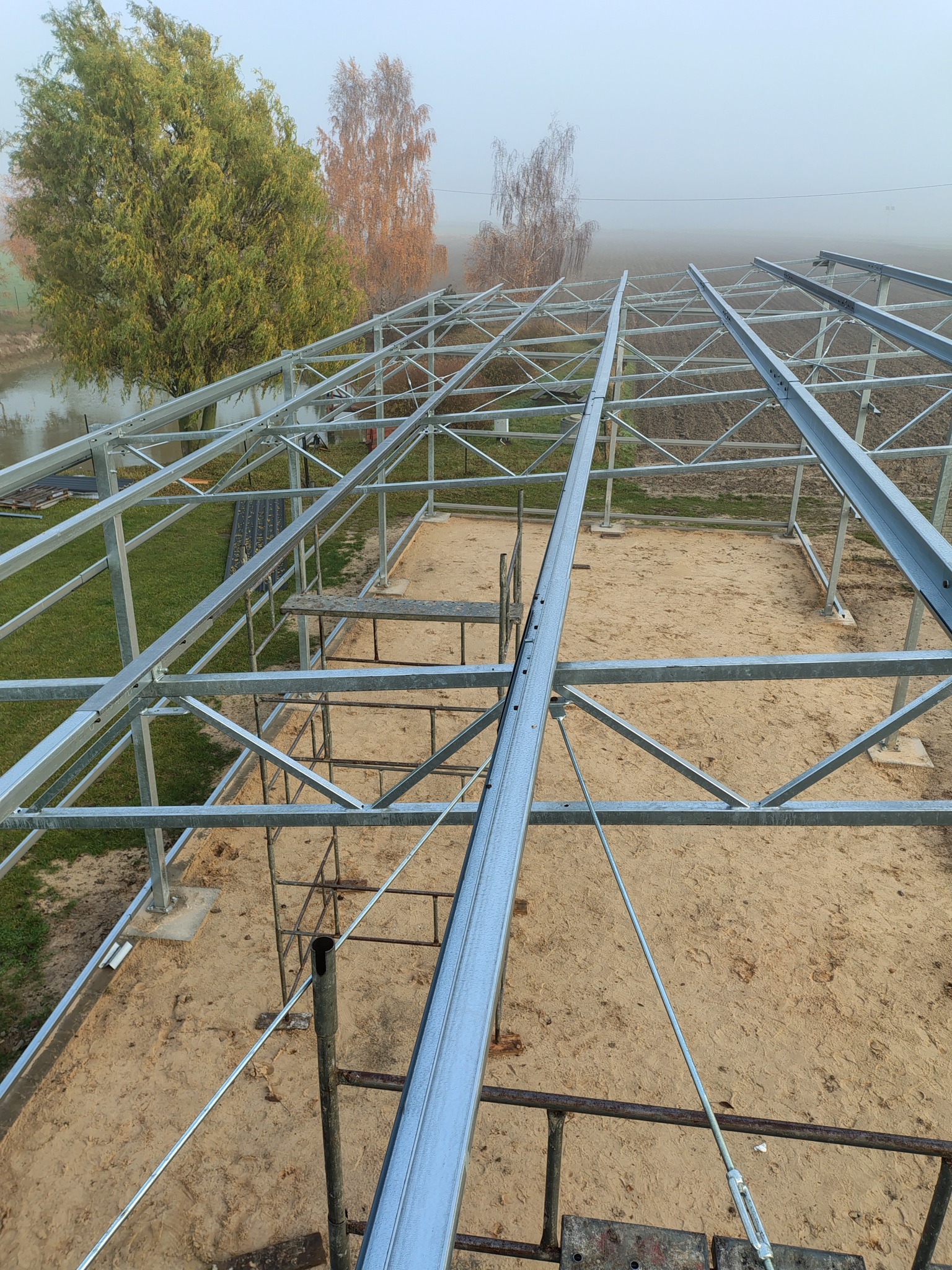 Metalowa konstrukcja szkieletowa budynku w trakcie budowy, widok z góry, mglista pogoda w tle. Elementy konstrukcyjne połączone śrubami.