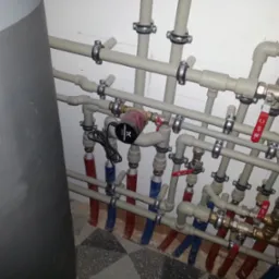 Rozbudowana instalacja grzewcza z rurami PEX w kolorach czerwonym i niebieskim, z pompą obiegową Grundfos i licznymi zaworami kulowymi, zamocowana do ściany za pomocą metalowych obejm.