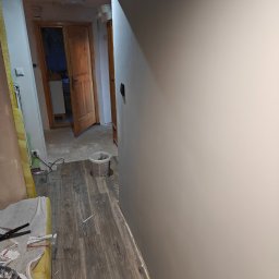 Przedsiębiorstwo Robót Budowlanych ART-DOM Artur Kuk - Korytarz w trakcie malowania: świeżo pomalowana ściana w odcieniach szarości, widoczne narzędzia malarskie i wiadro z farbą, drewnopodobna podłoga. W tle otwarte drzwi.