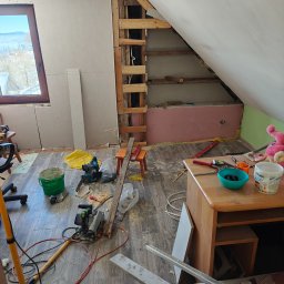 Przedsiębiorstwo Robót Budowlanych ART-DOM Artur Kuk - Poddasze w trakcie adaptacji: widoczne płyty karton-gips, drewniana konstrukcja, narzędzia i bałagan. Okno z widokiem na zimowy krajobraz.