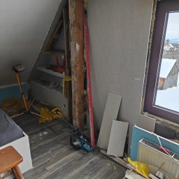 Przedsiębiorstwo Robót Budowlanych ART-DOM Artur Kuk - Poddasze w trakcie remontu: odsłonięta konstrukcja, płyty karton-gips, narzędzia i lampa budowlana. Widok na zaśnieżony krajobraz przez okno.