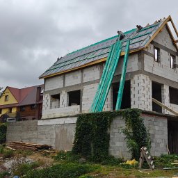 Przedsiębiorstwo Robót Budowlanych ART-DOM Artur Kuk - Budowa domu z białych bloczków: ekipa montuje izolację dachu, zielone deski opierają się o ścianę, w tle sąsiedni dom i pochmurne niebo.