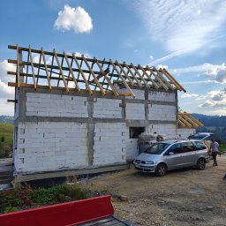 Przedsiębiorstwo Robót Budowlanych ART-DOM Artur Kuk - Budowa domu z białych bloczków, widoczne drewniane elementy konstrukcji dachu, auto dostawcze z materiałami budowlanymi w tle, krajobraz górski.