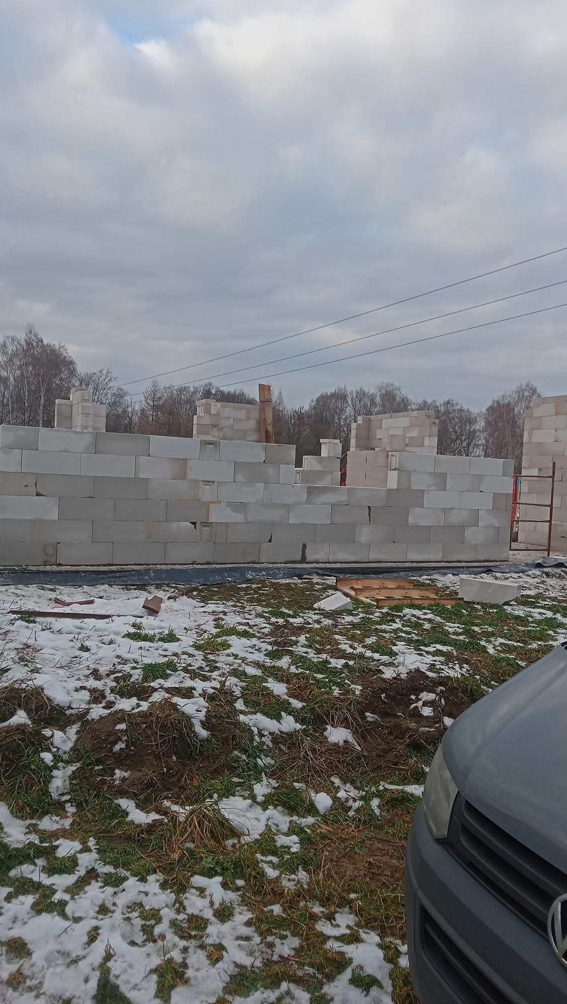 Wznoszenie ściany z białych bloczków betonu komórkowego na fundamencie. Częściowo ośnieżony teren budowy, pochmurne niebo. Widoczny fragment samochodu.