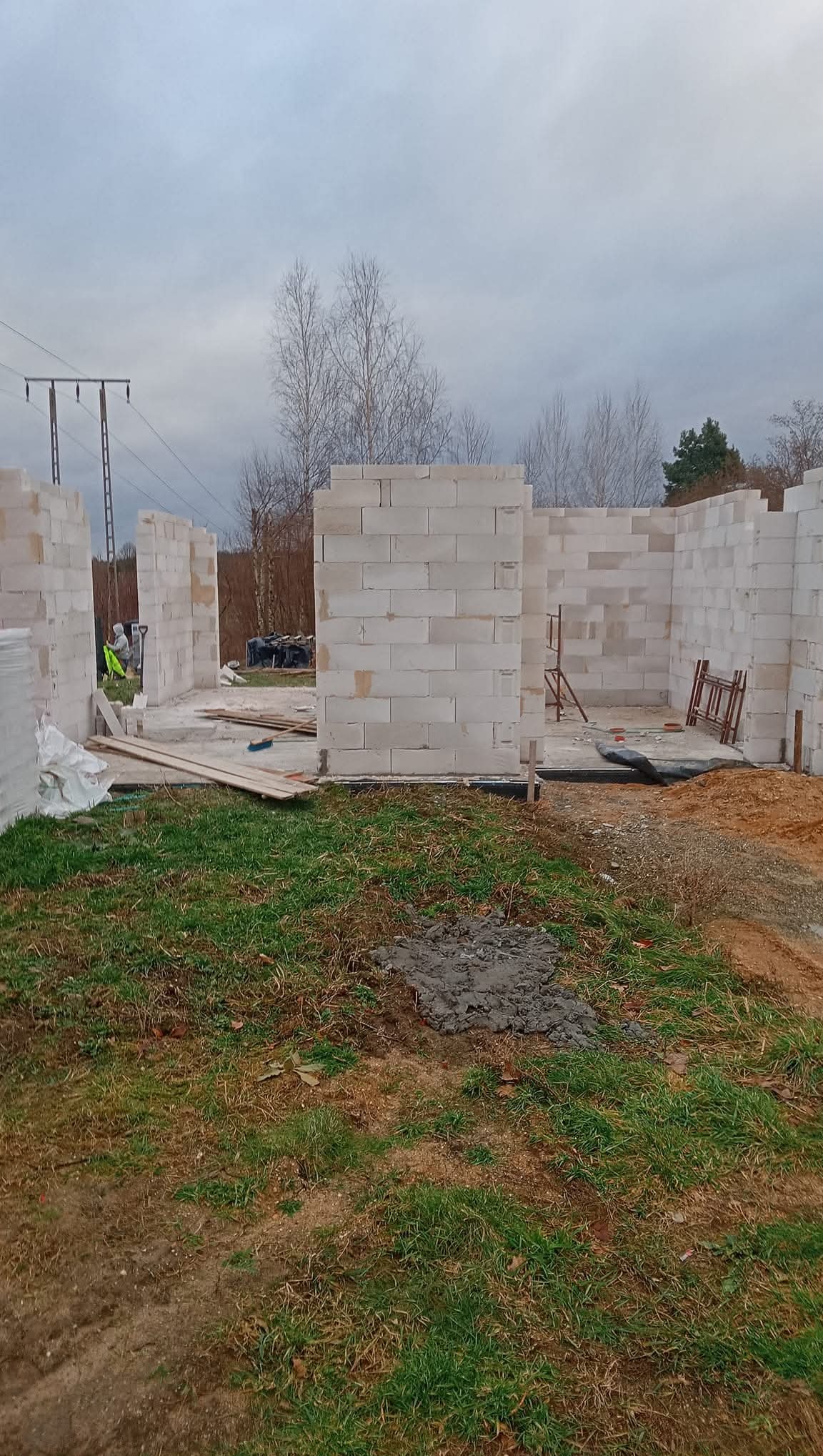 Wznoszenie ścian parteru domu z bloczków betonu komórkowego. Widoczne fragmenty ścian, materiały budowlane i teren budowy w pochmurny dzień.