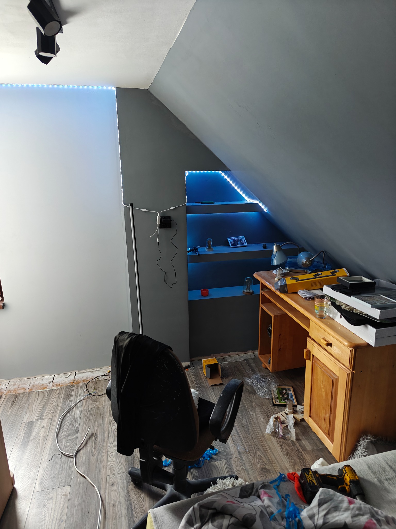 Pokój na poddaszu w trakcie remontu: szare ściany, drewniana podłoga, biurko z narzędziami, oświetlone półki wnękowe. Widoczne kable i ślady prac remontowych.